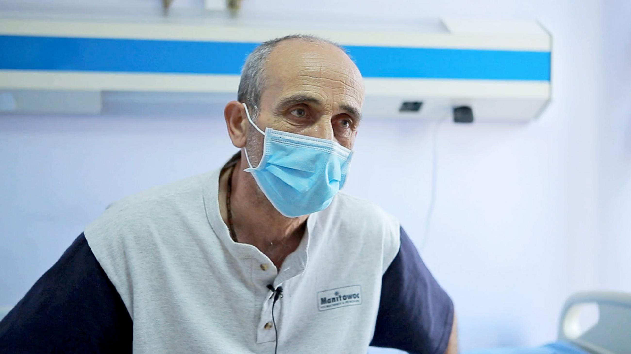 A middle-aged man wearing a light-blue surgical mask sits in a doctor's office. 