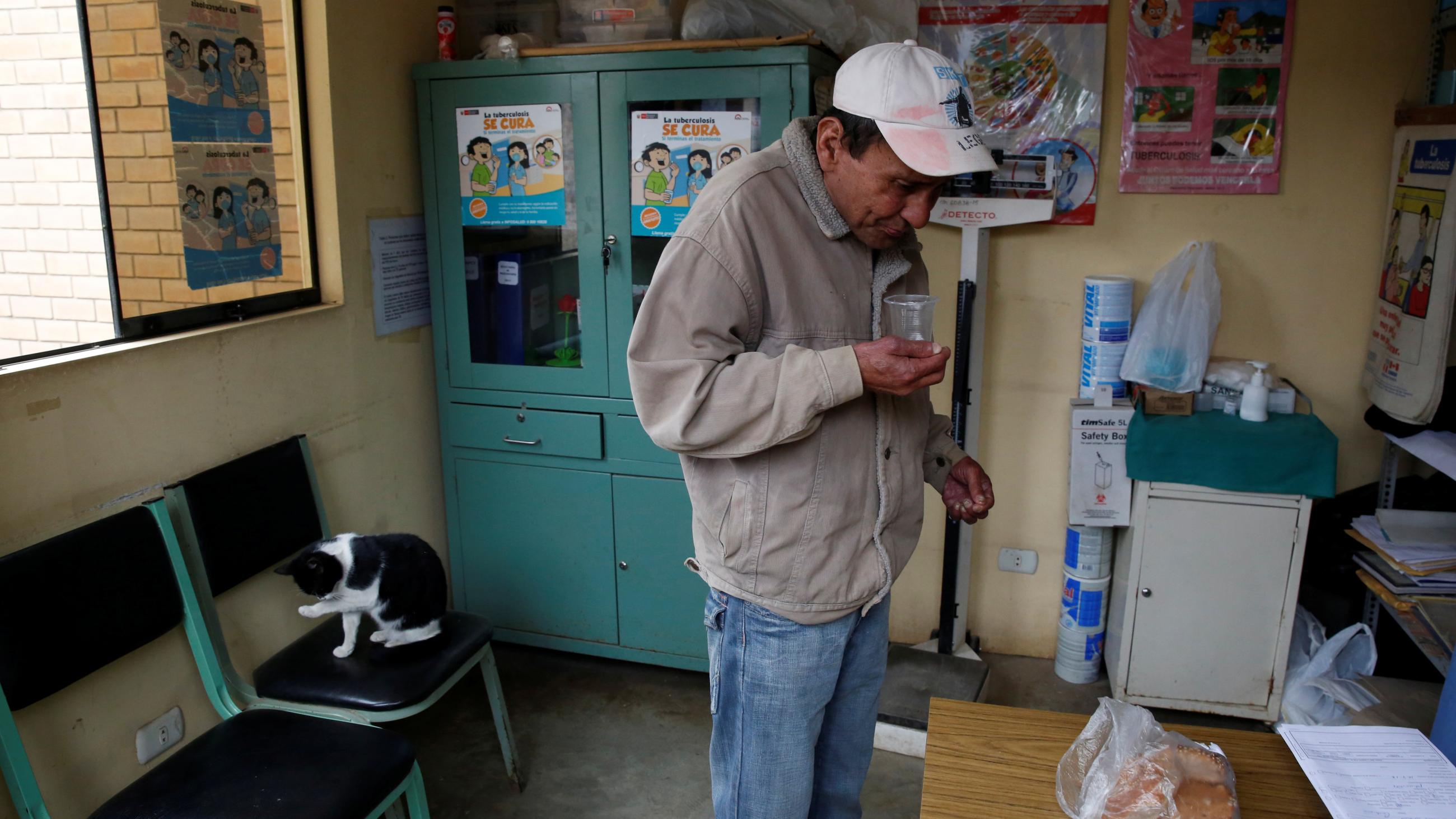 A man wearing a khaki jacket and ball cap takes medication with a glass of water.