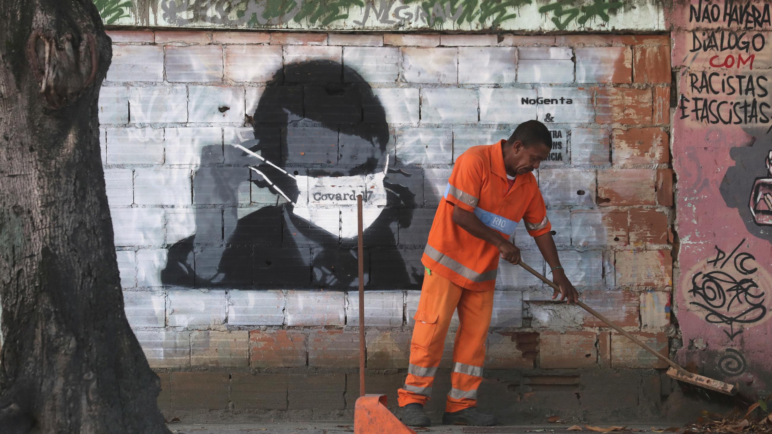 A municipal worker cleans a street in front of street art depicting Brazilian President Jair Bolsonaro adjusting his face mask during the COVID-19 outbreak in Rio de Janeiro, Brazil, on March 26, 2020.