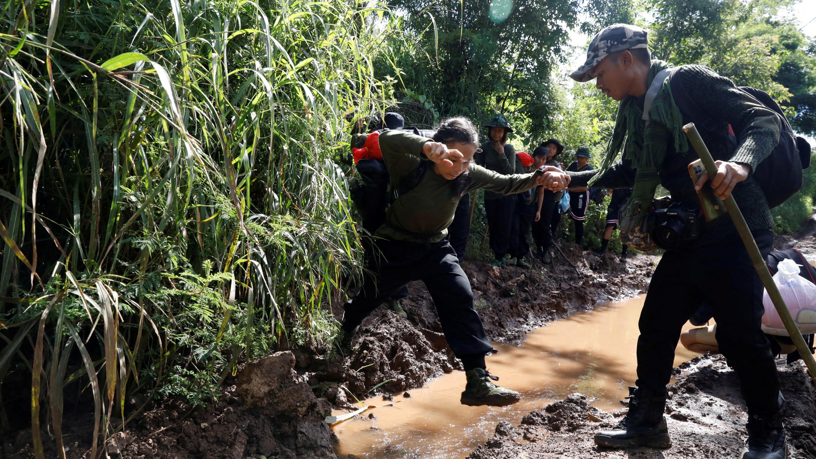 A rebel soldier helps a young female soldier cross a muddy stream surrounded by dense green jungle.