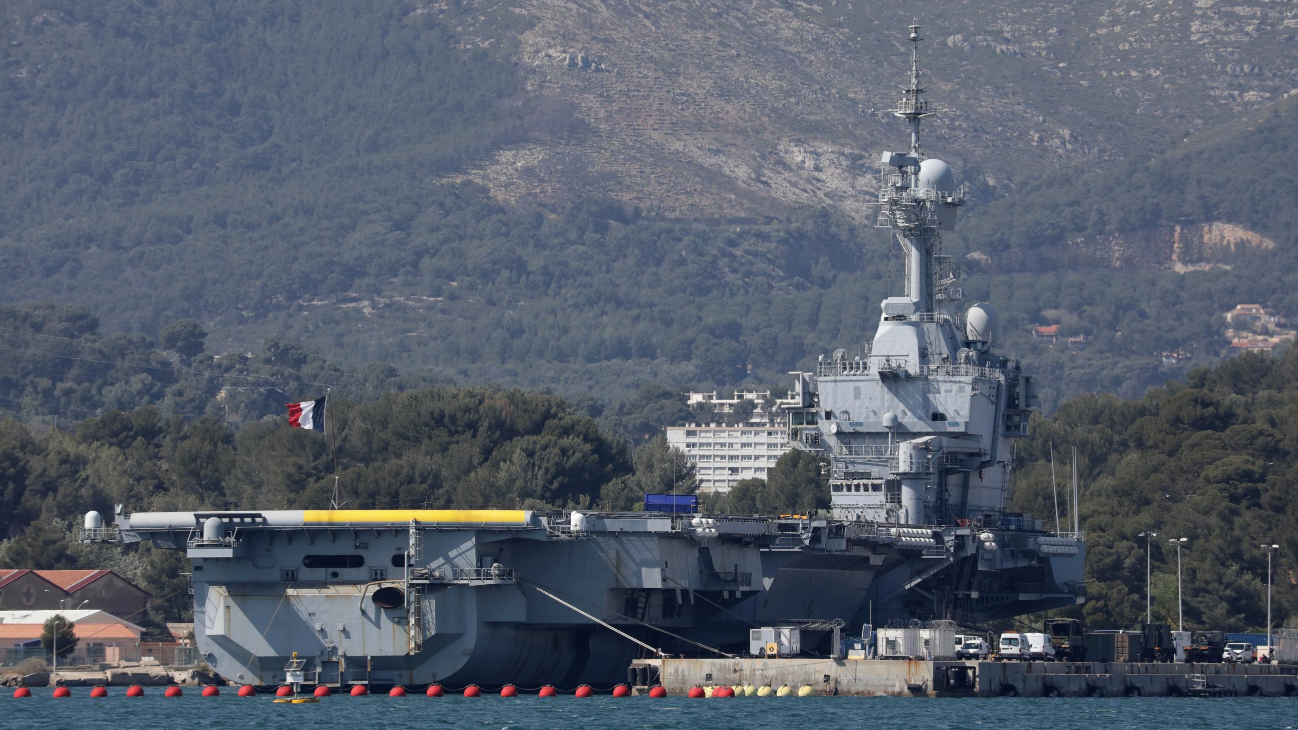 An aircraft carrier waving the French tricolor flag is docked at a port against a backdrop of mountains.