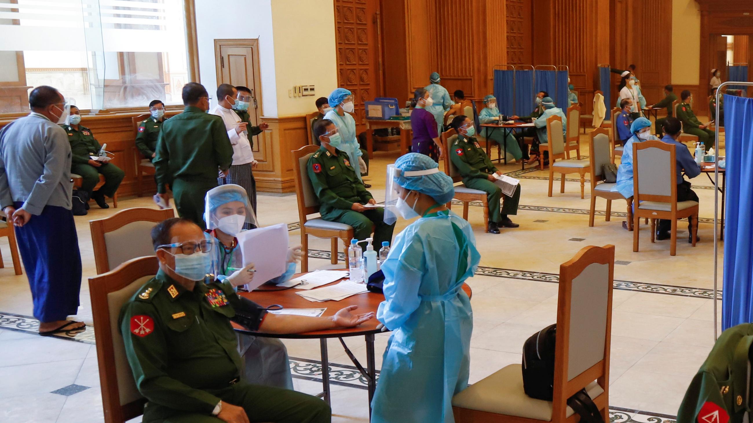 Nurses in turquoise medical gowns administer vaccines to military officers in green uniforms bedecked with medals.