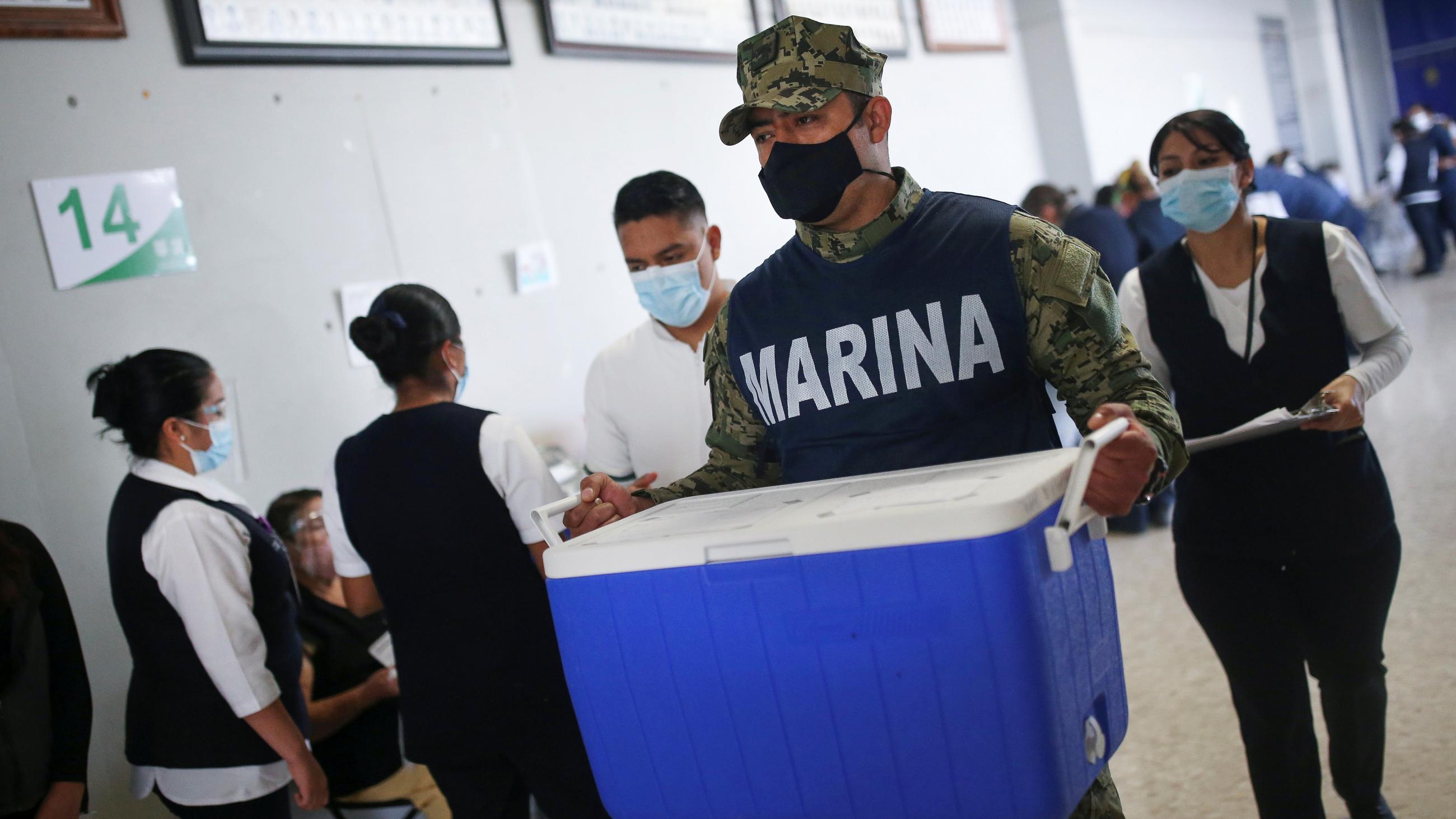 A soldier in a navy blue Marine Corps vest carries a blue cooler of vaccines through an active vaccination site.