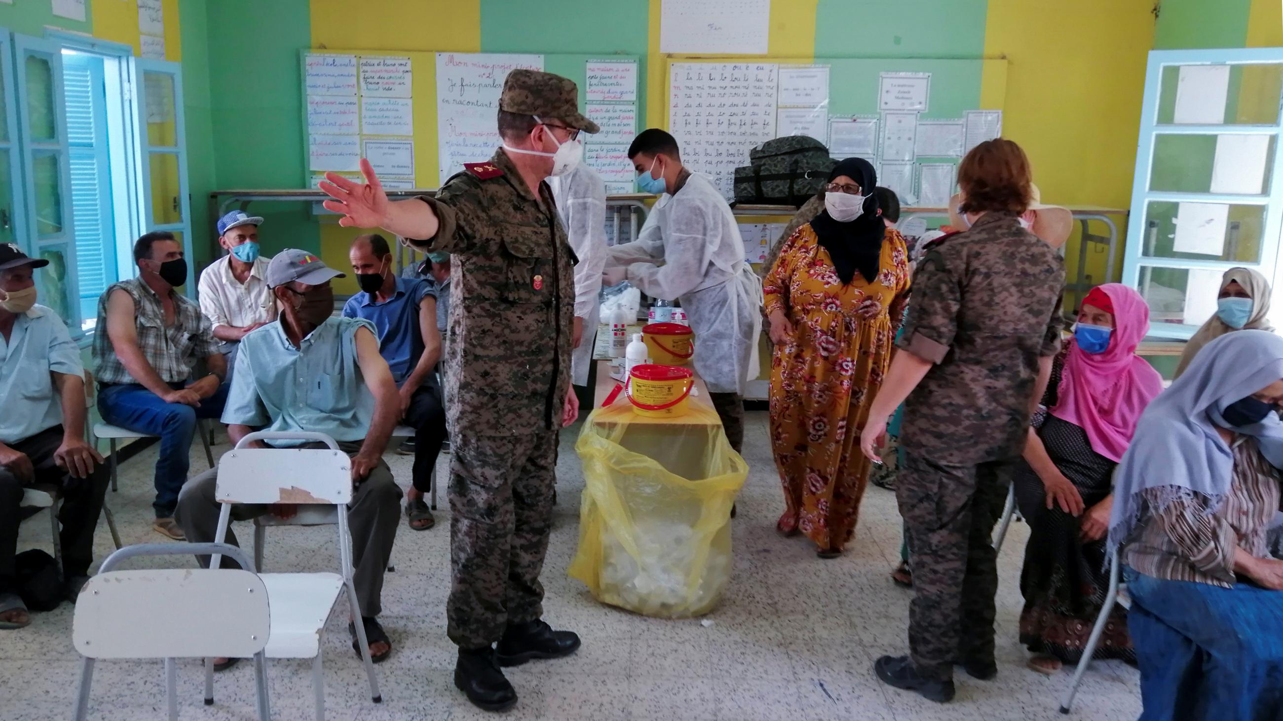 A soldier directs women in colorful dresses and headscarves in a classroom that doubles as a vaccination site.