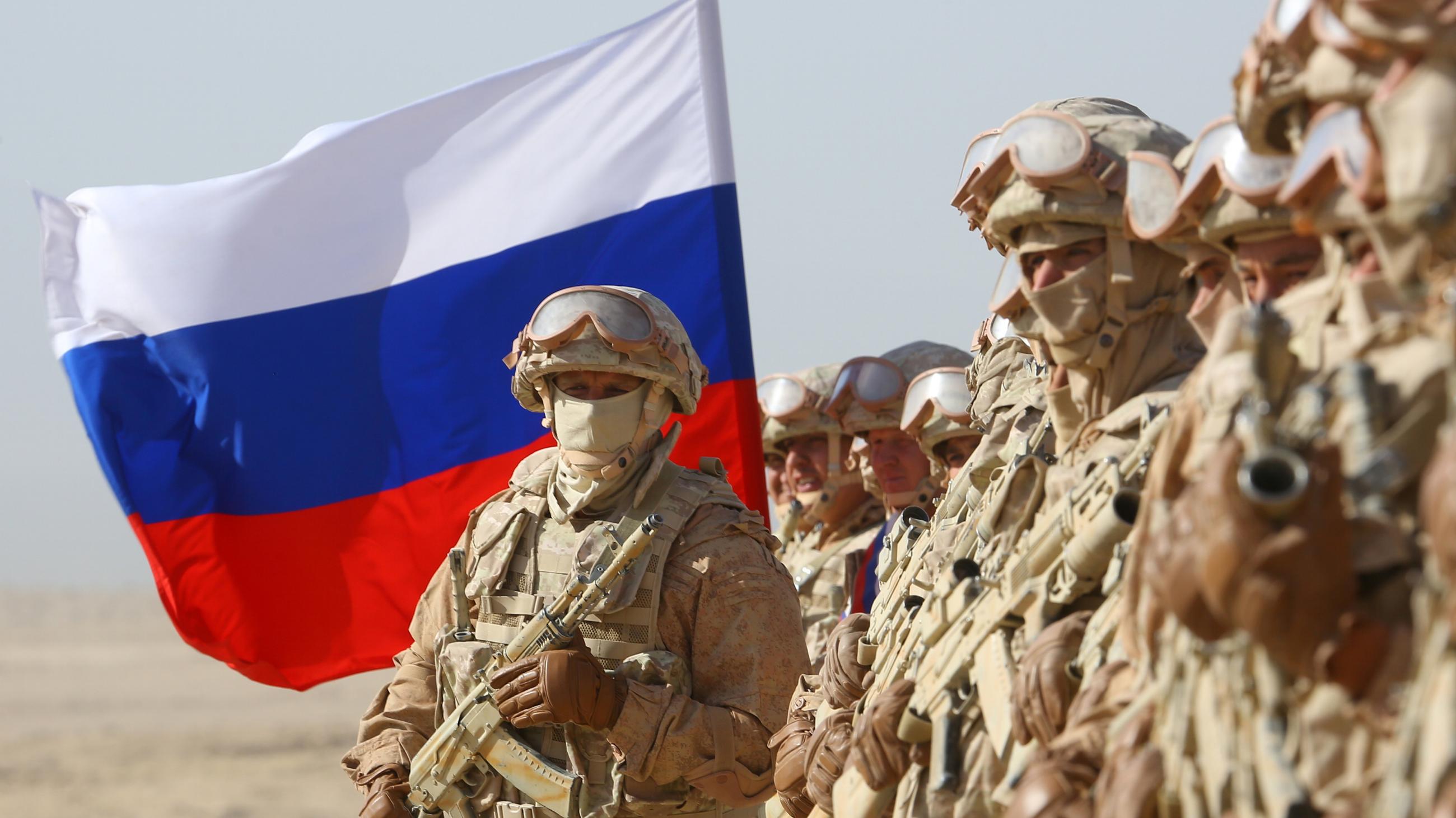 Russian soldiers in khaki-colored combat gear bearing rifles stand in a line in the desert with a Russian flag.