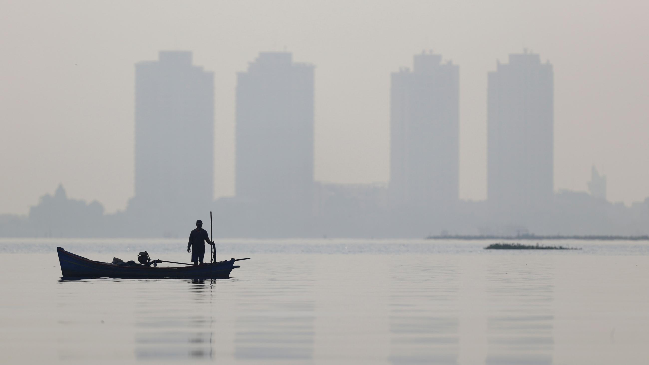 A fisherman stands on a wooden boat as smog covers high rise buildings seen in the background, at the north coast of Jakarta, Indonesia, on August 28, 2021. 