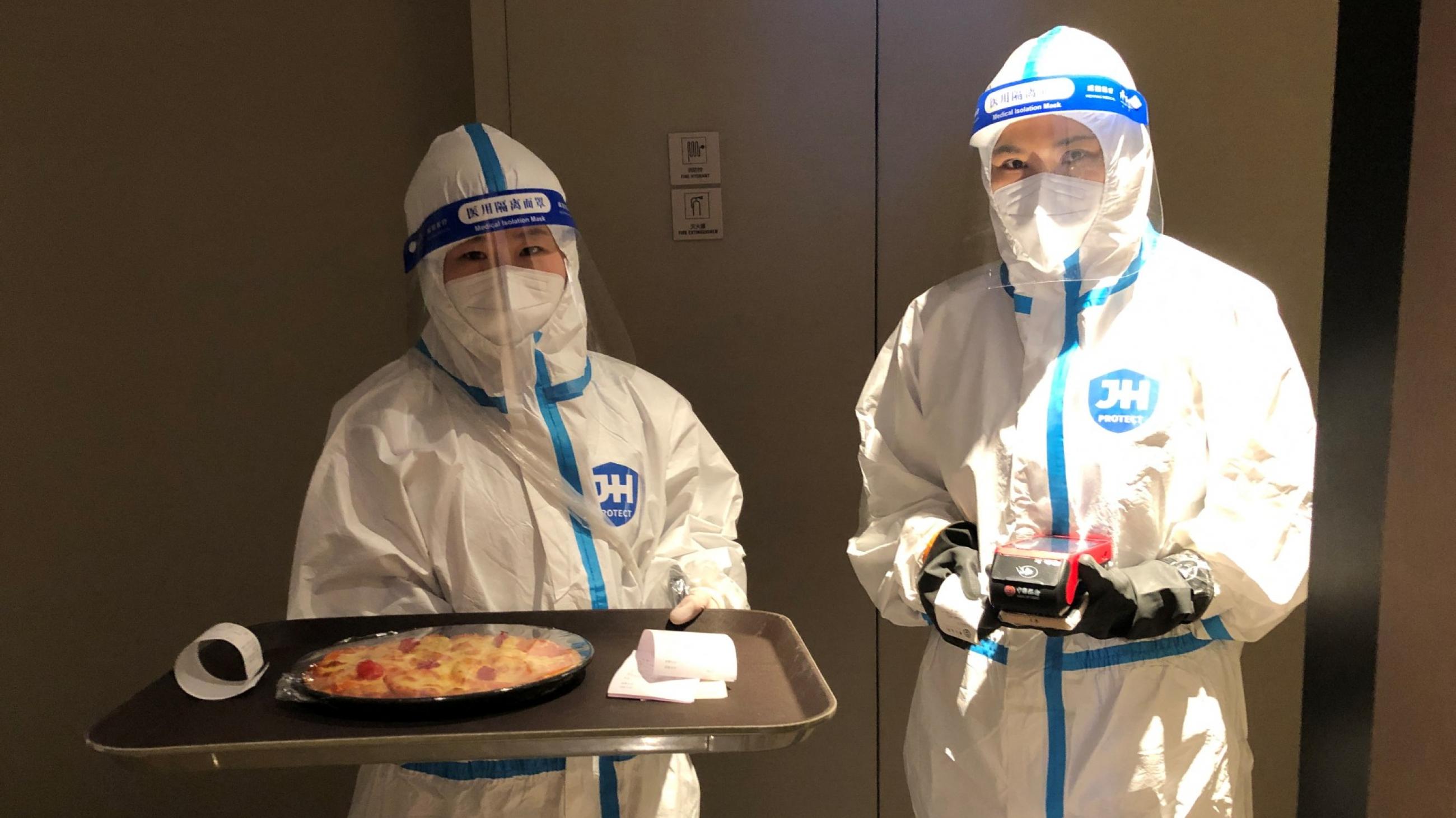 Two hotel staff wearing personal protective equipment (PPE) deliver a Margherita pizza wrapped in cling film that was ordered with room service at a hotel for journalists and officials o
