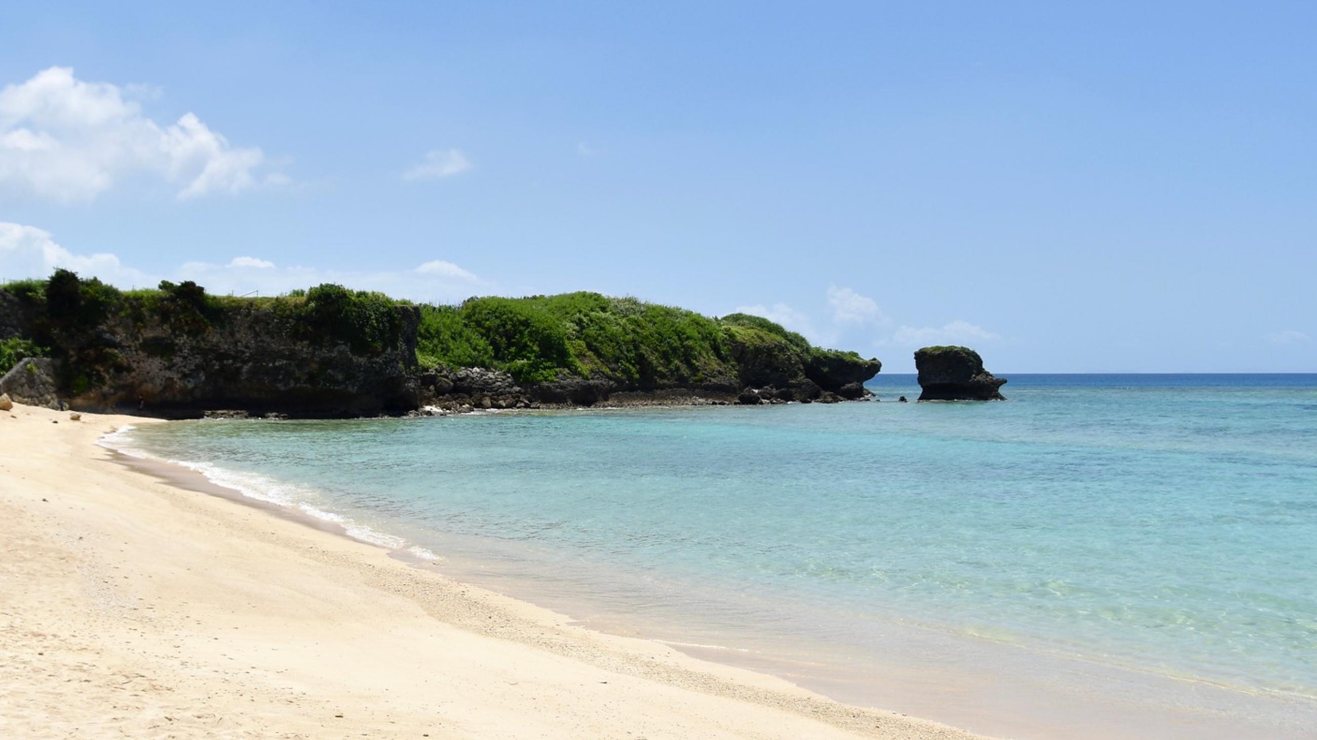 A golden sandy shoreline with light crystal blue waters is captured in Okinawa, Japan