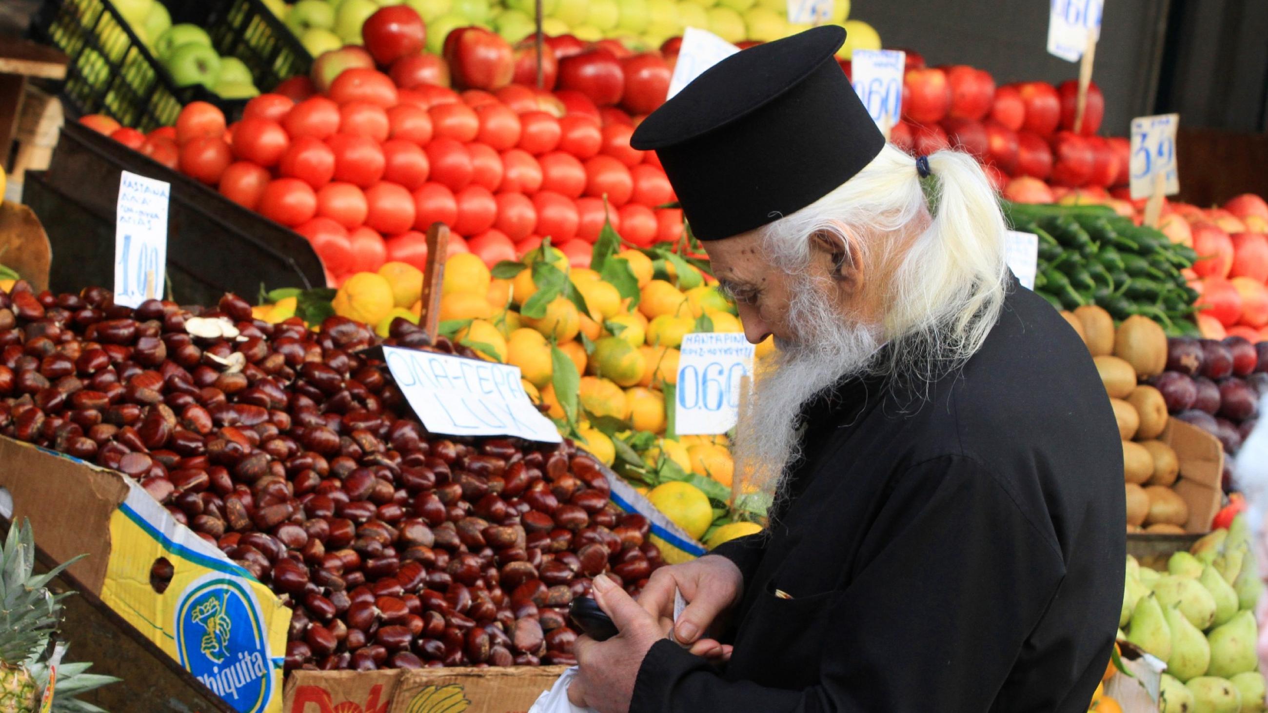 An orthodox priest with a long white beard wearing traditional black robes and hat is seen examing fresh fruits and vegetables at a market stall
