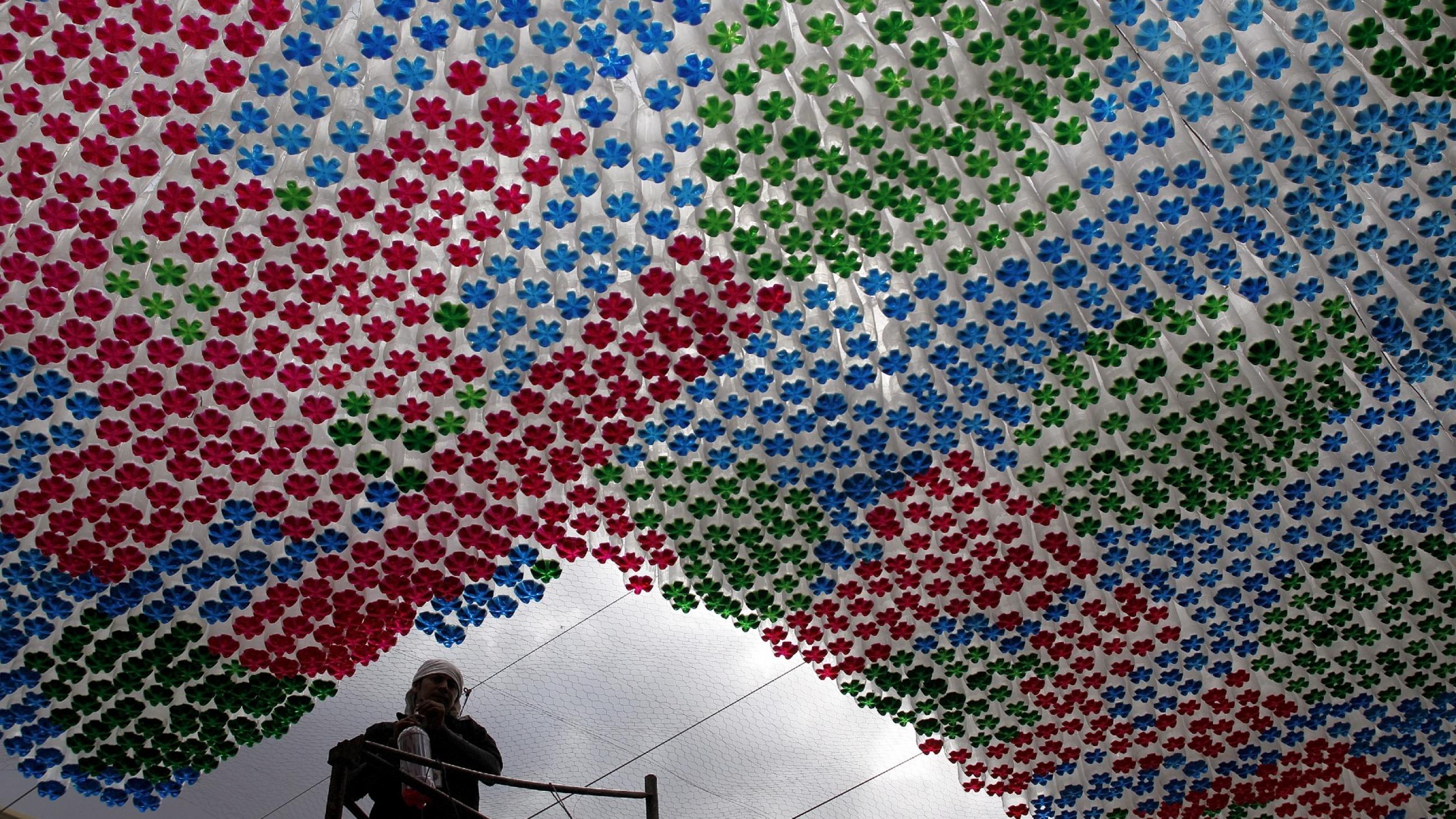 a museum worker, obscured by light, cleans next to an art installation made from repurposed, red, blue, and green bottles