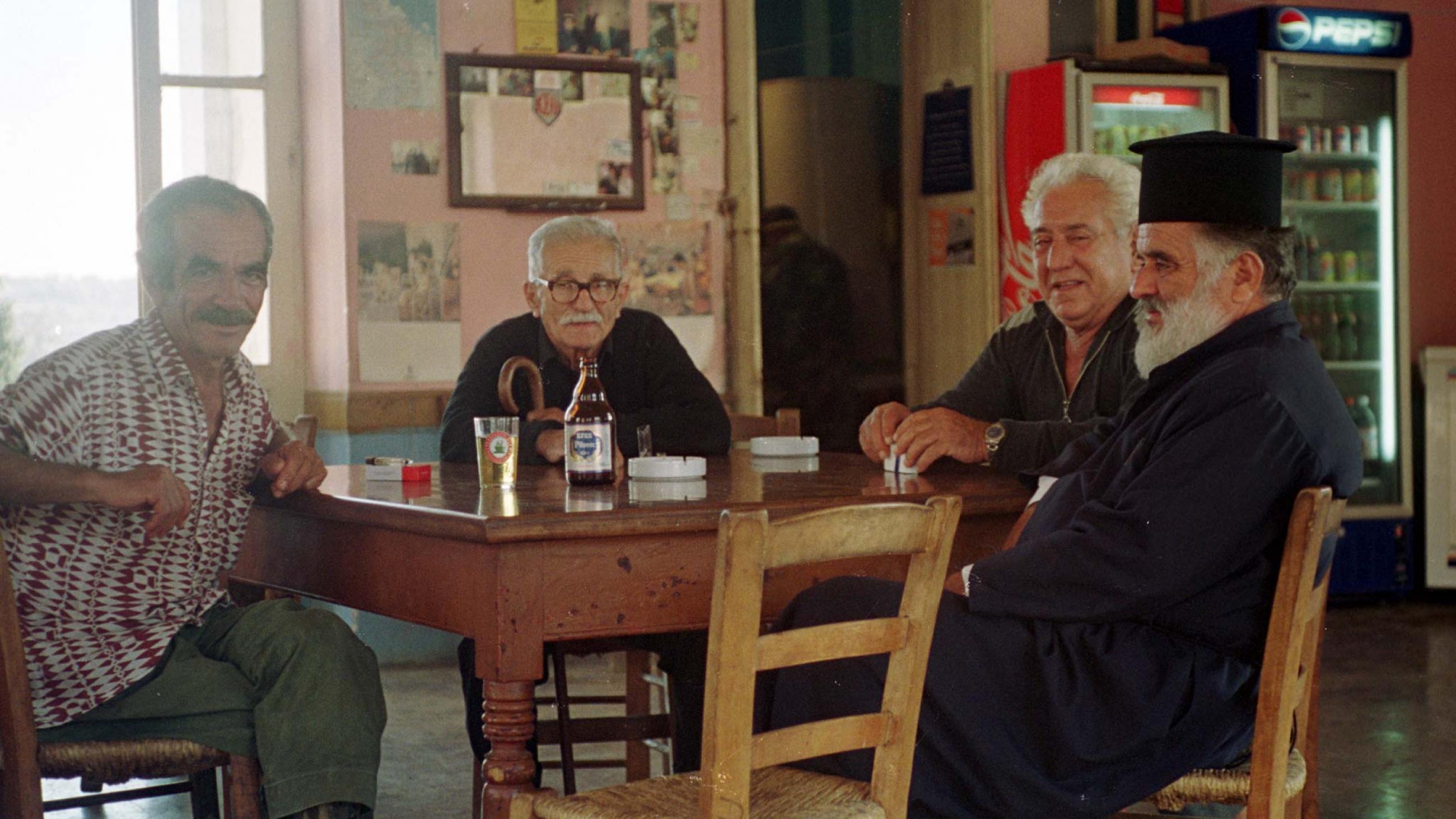 a group of four elderly greek men sit around a wooden table at a small village cafe, From left to right: the first man is wearing a white and red checked shirt with jeans, the next two are wearing black half zip shirts, the fourth man is an Orthodox priest in traditional garments