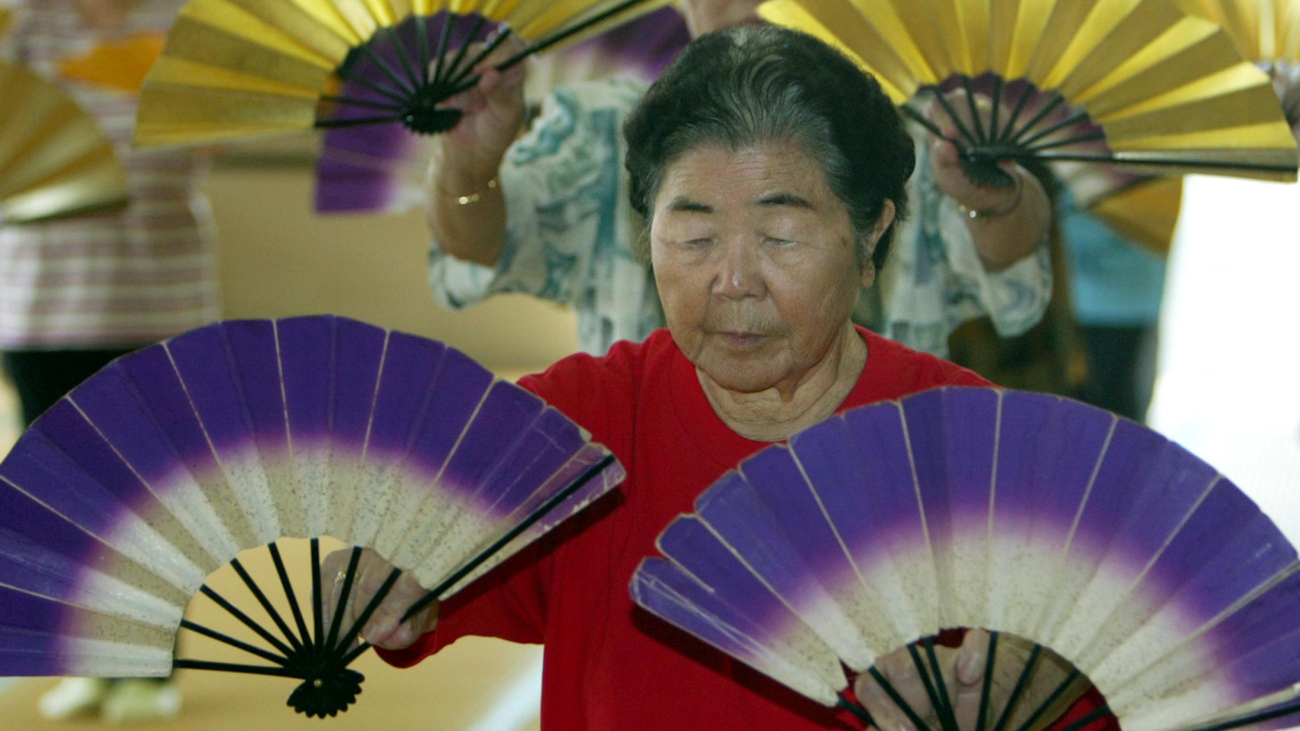 An elderly japanese woman wearing a red t shirt dances holding white and purple fans, in a group