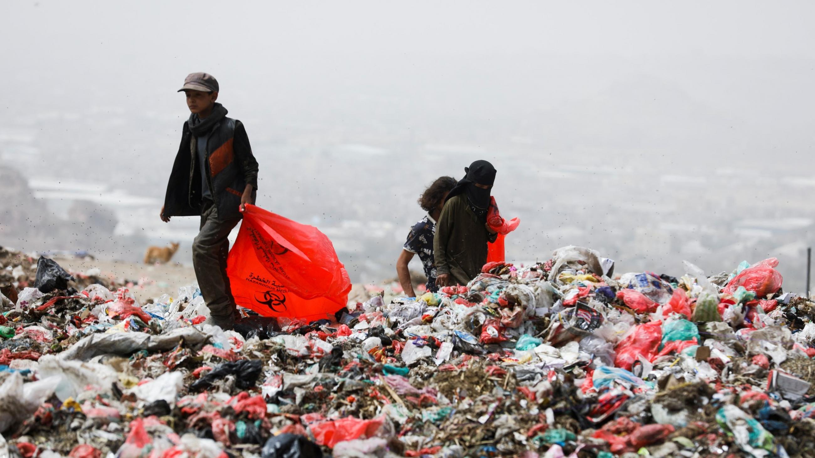 Two men wearing black jackets and pants carrying red plastic bags search a landfill for recyclable plastics