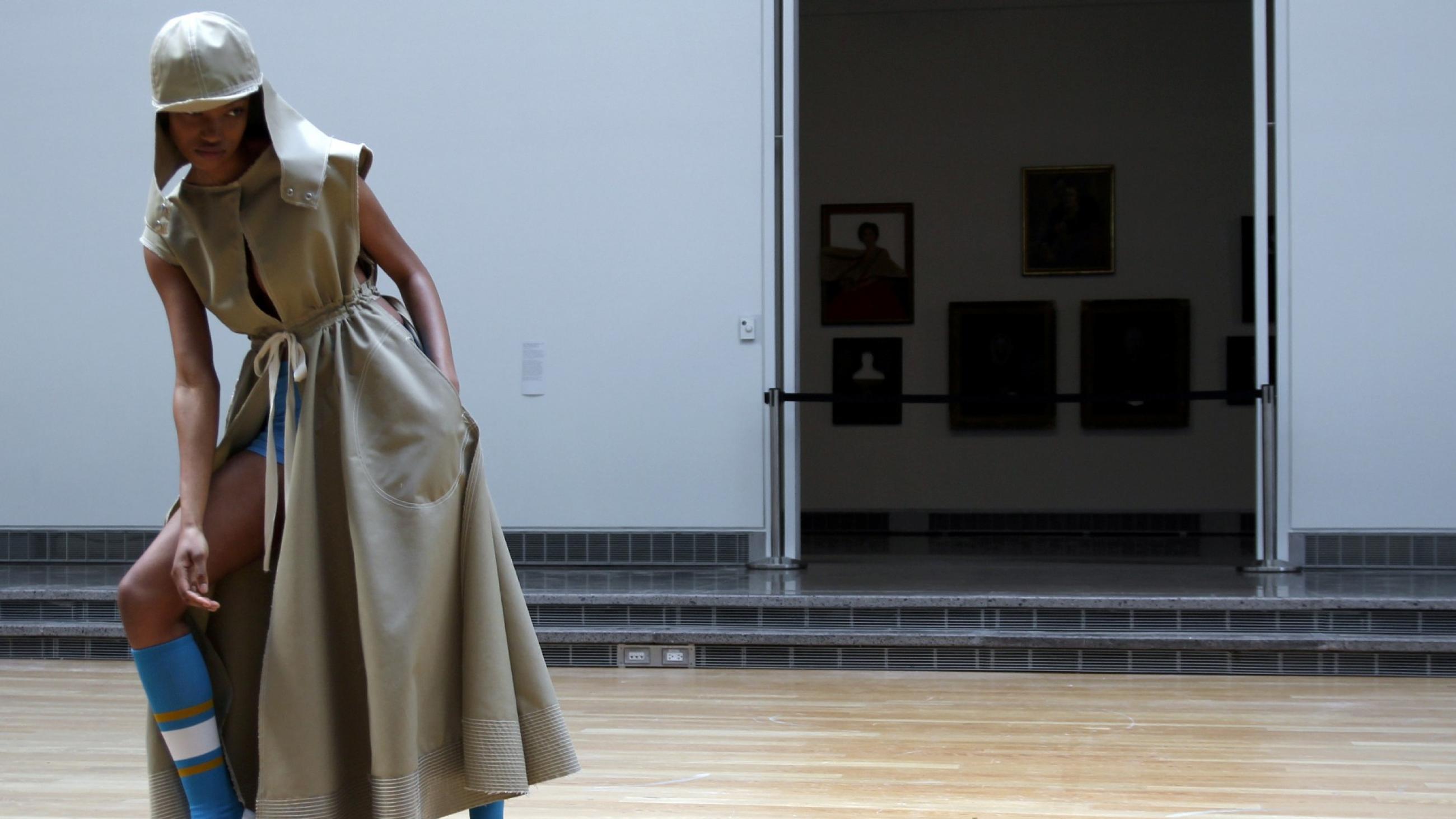 A model, wearing a khaki colored sleeveless trench coat made from recycled plastics, and blue knee high socks, strikes a pose in a dance hall as part of an art installation