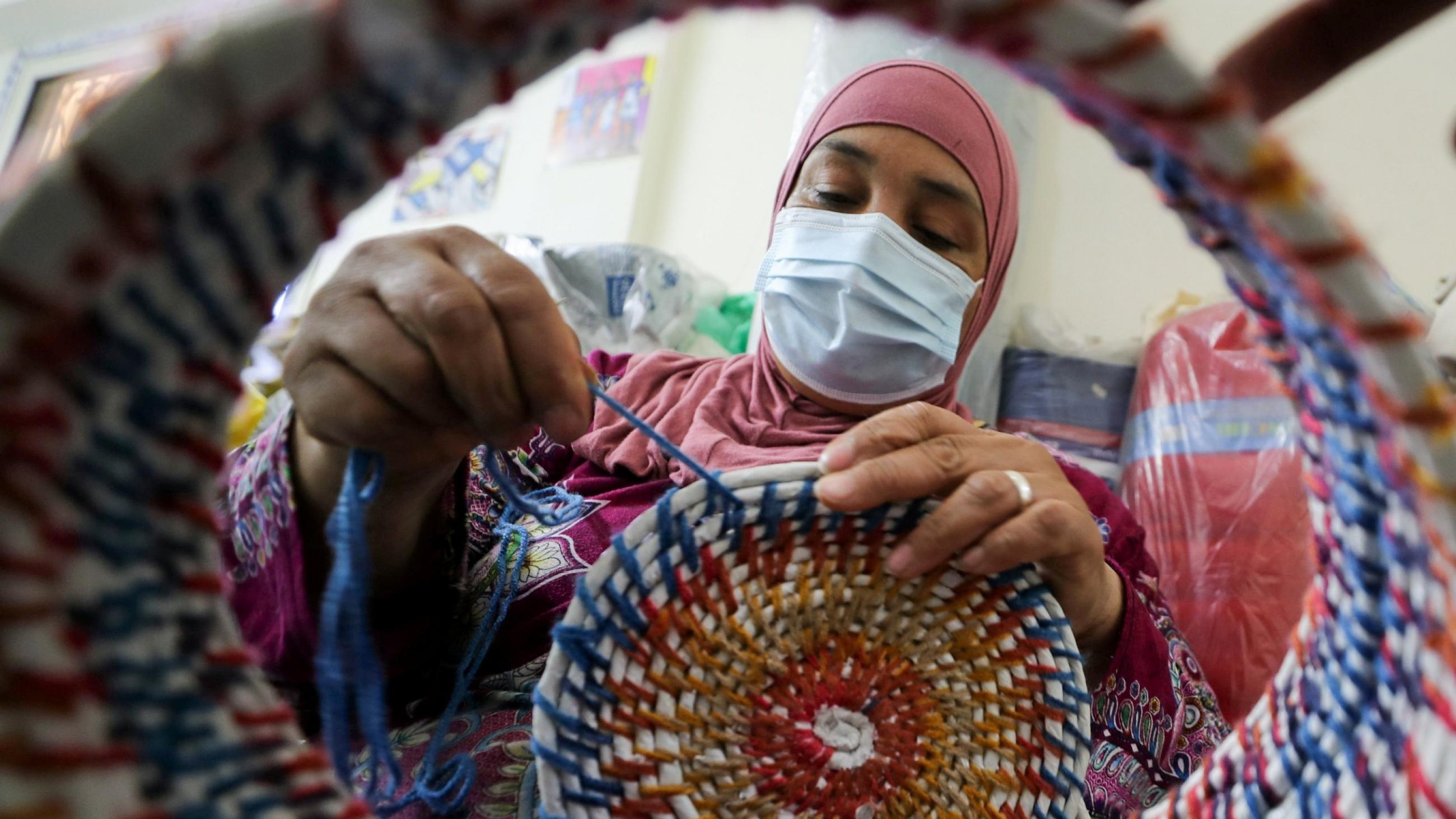 An Egyption woman wearing a pink headscarf a blue surgical mask is seen knitting a colorful bowl out of yarn made form recycled plastics