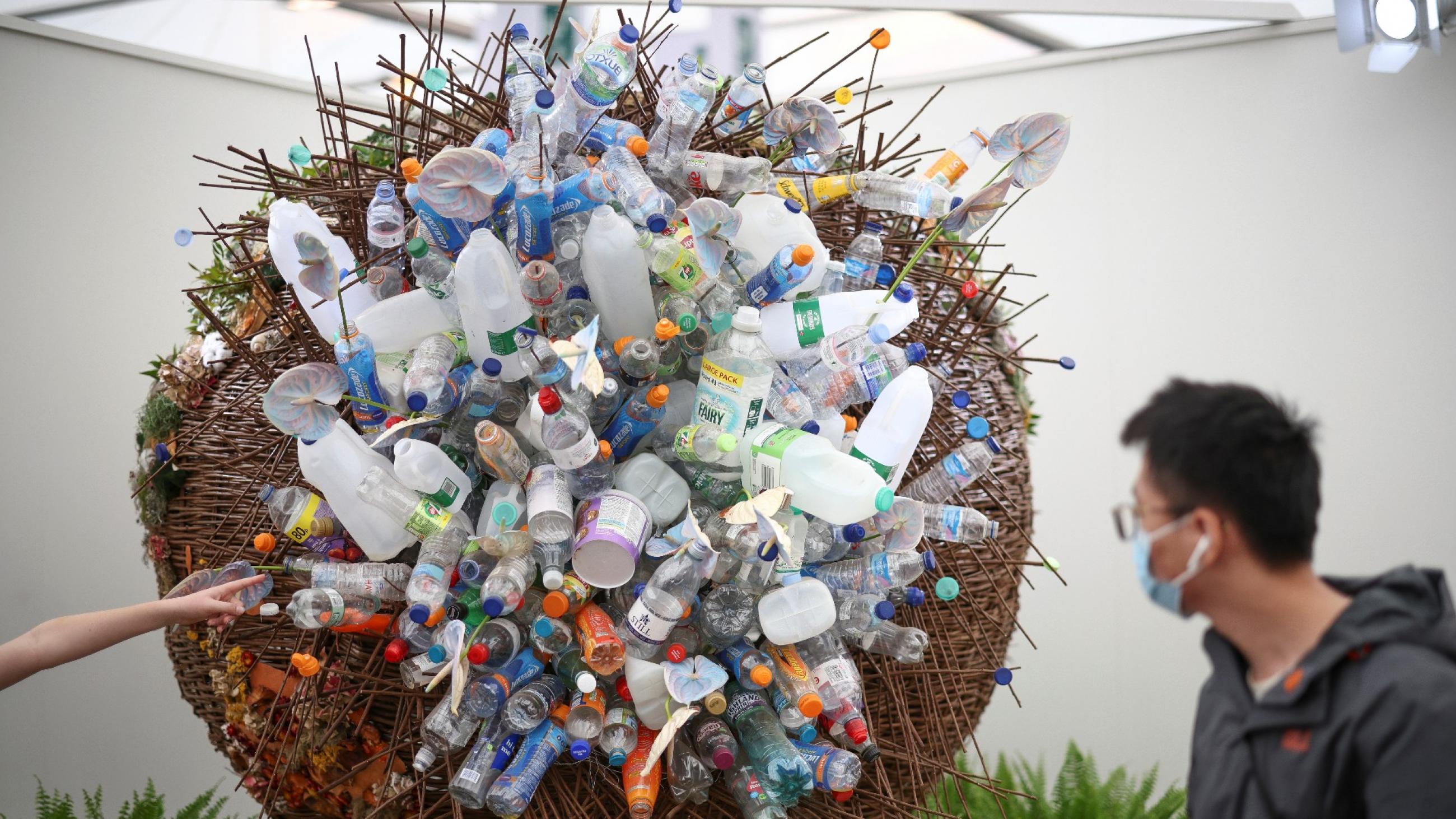 A man with black hair wearing a blue surgical face mask looks at a sculpture made out of plastic bottles at he Chelsea Flower Show, in London, England