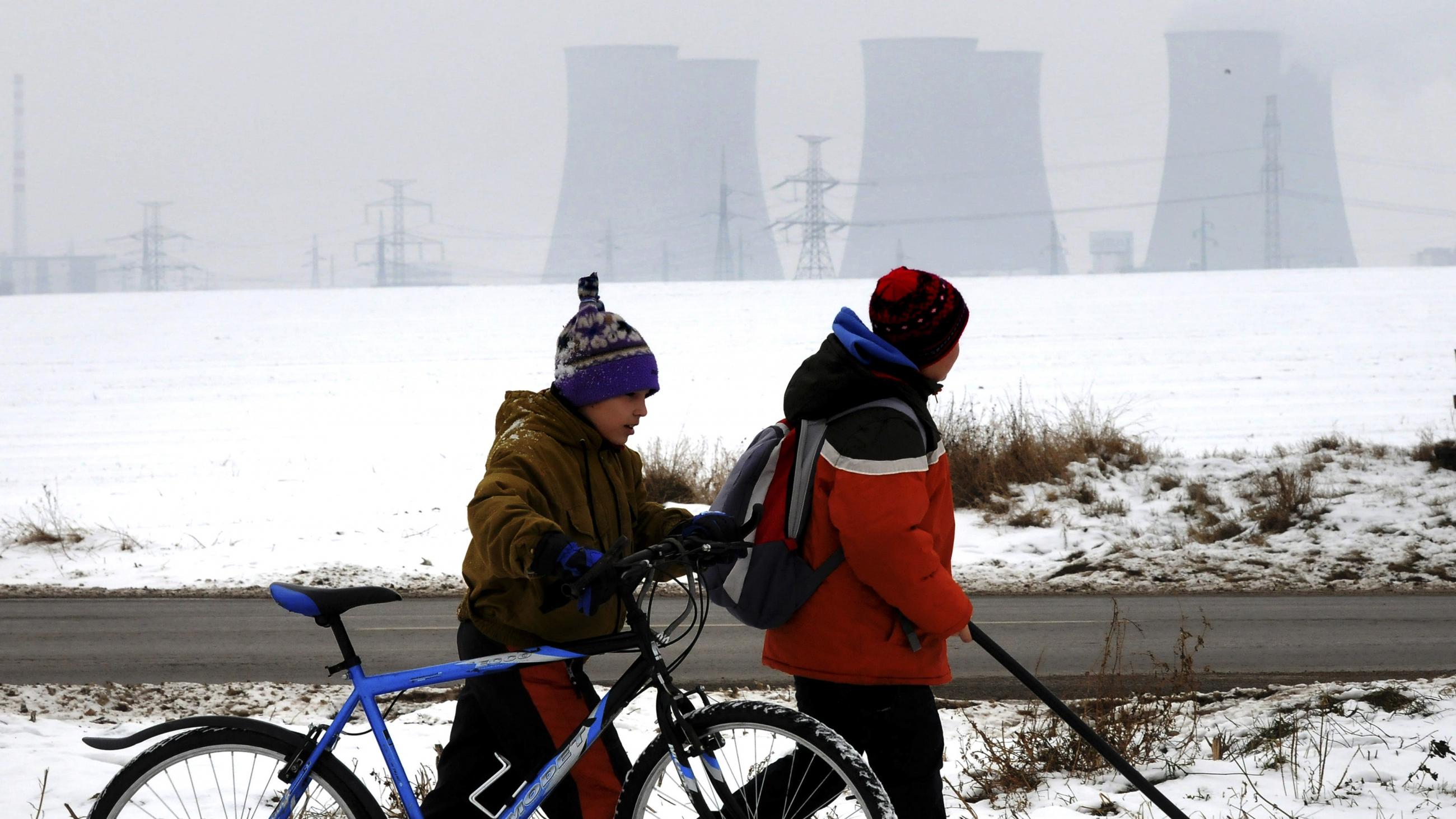Two young boys, on in a red coat with a hockey stick and one in a brown coat with a blue bicycle walk along a snowy path next to a river. On the other side of the river, shrouded in fog are six cooling towers of a nuclear power plant