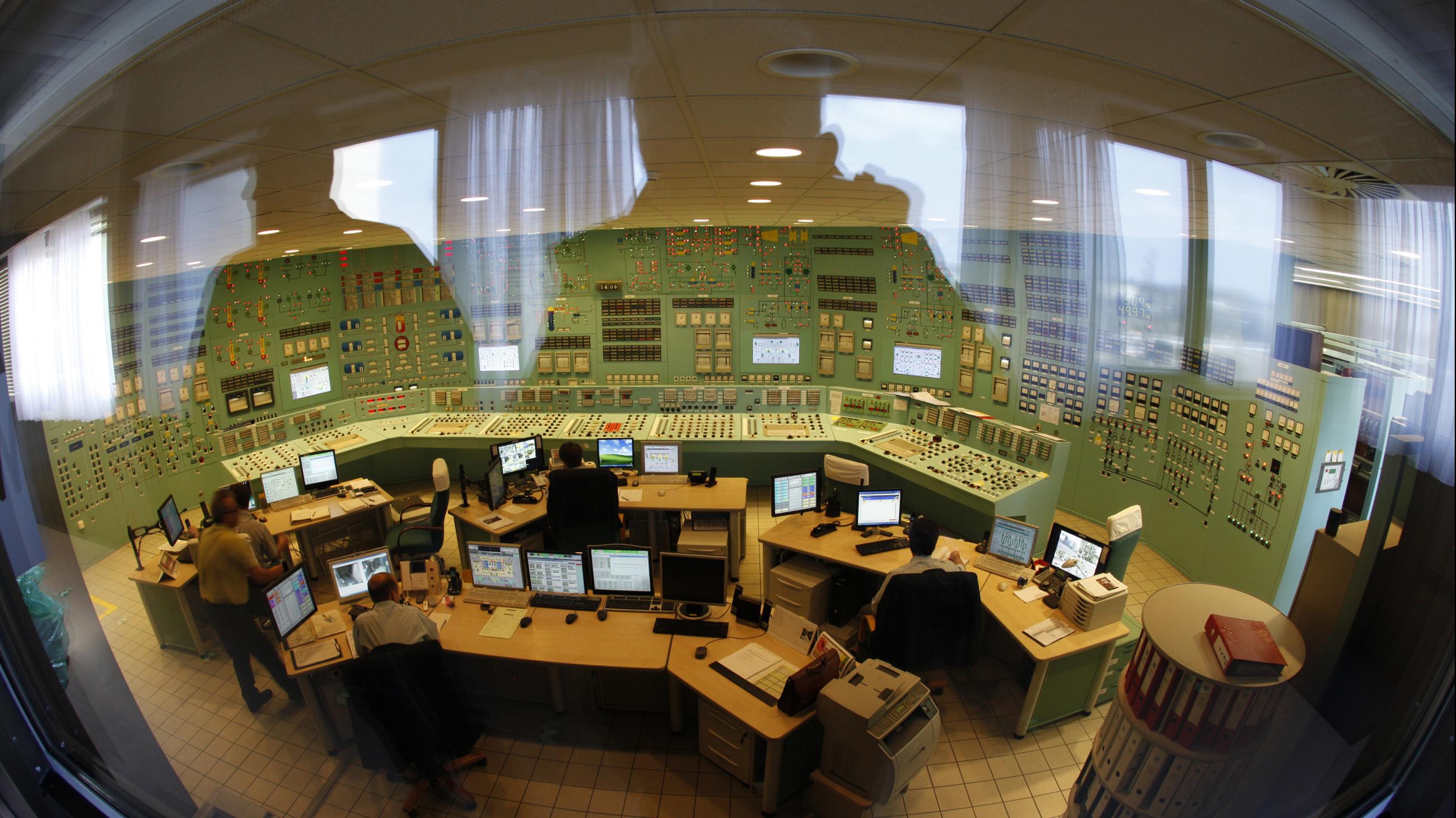 Through a large glass window, the viewer can see the silhouetted reflection of the photographer and another figure superimposed over nuclear power plant workers sitting in front of computer screens in a mint green control room the walls of which are covered in dials and knobs