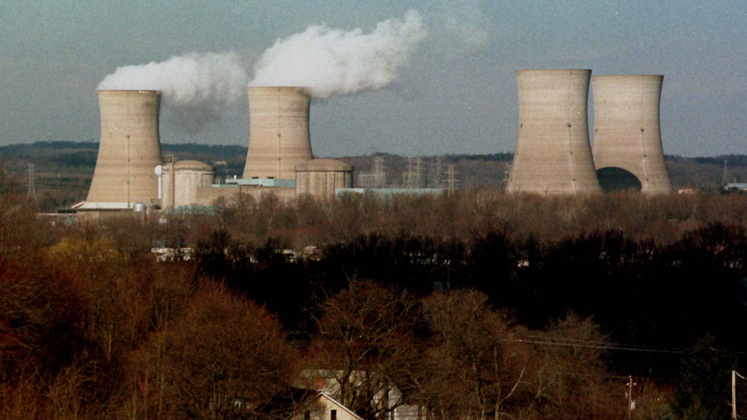 Four nuclear reactors, two dormant and two with columns of white smoke, stand against a blue sky. In the foreground are the bare branches of a forest in winter surrounding some small light-colored houses