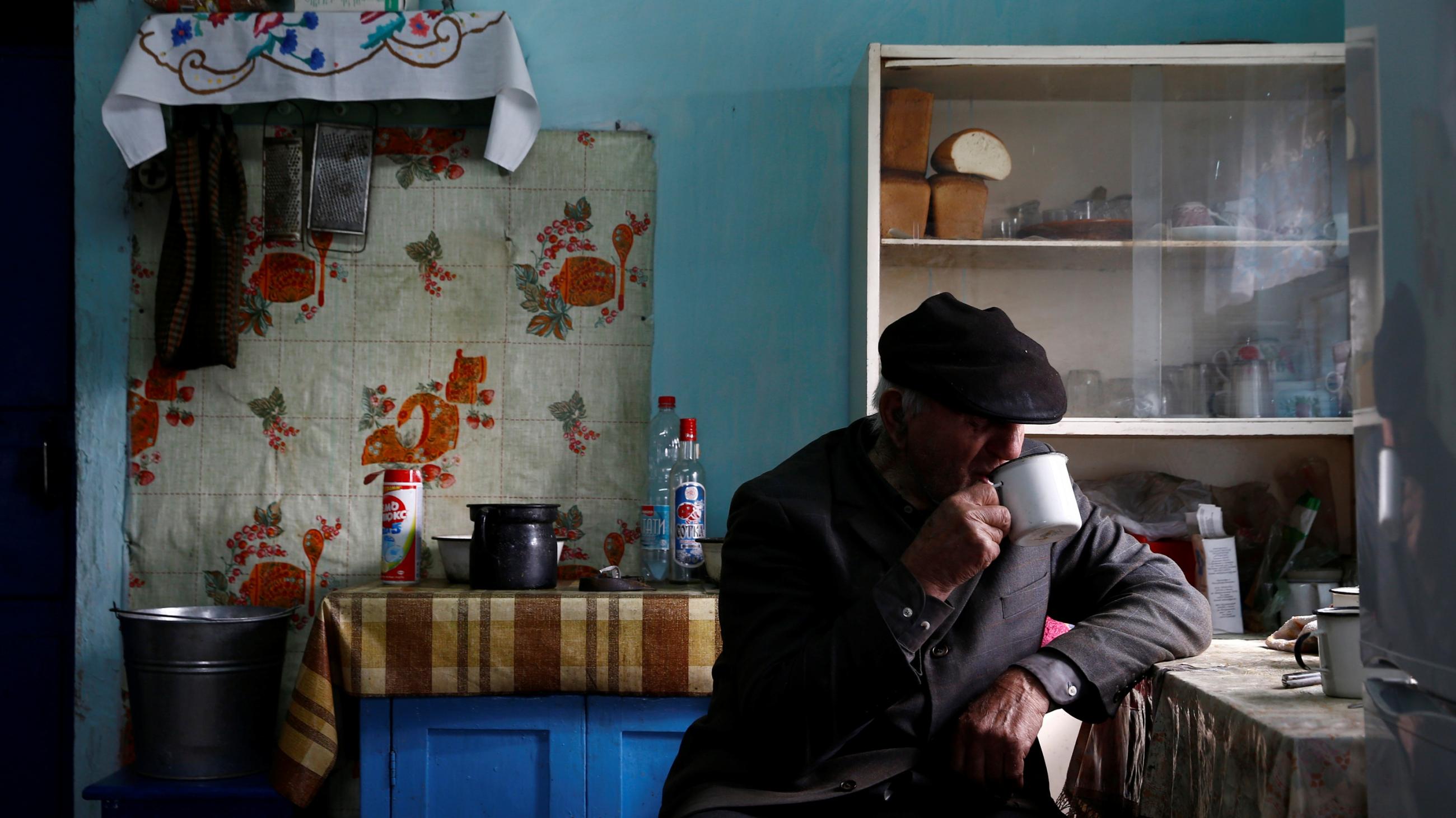 An old Belarusian man in a black hat drinks tea in his Soviet-style kitchen, which has turquoise walls, shelves with bread, and white tiles with red flowers above a small stove. It looks as though it hasn't changed since the 1980s.