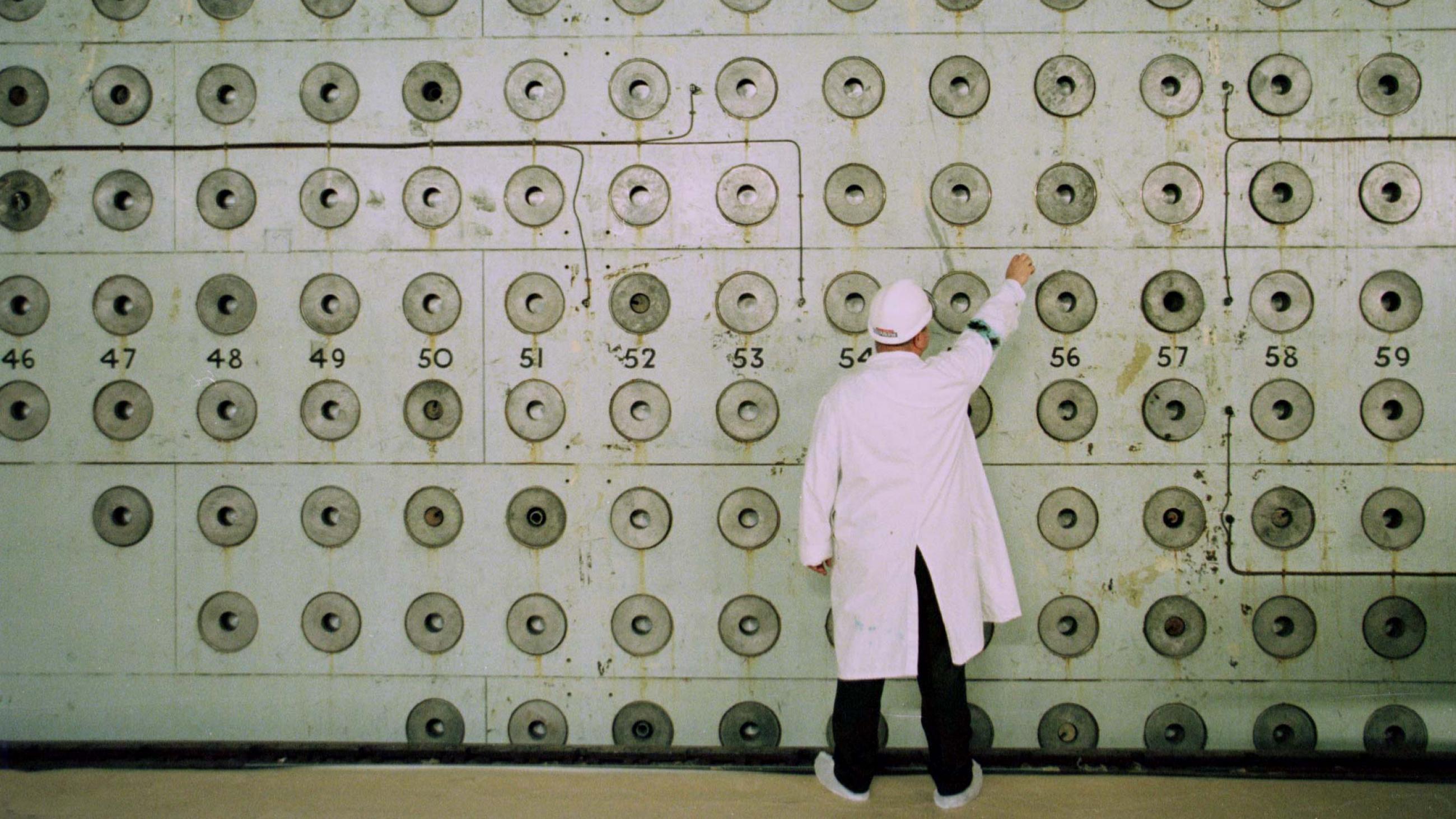 A nuclear plant worker in a white lab coat and medical hat adjust knobs on a pale green wall covered with number dials at the Windscale Pile One nuclear power plant. 