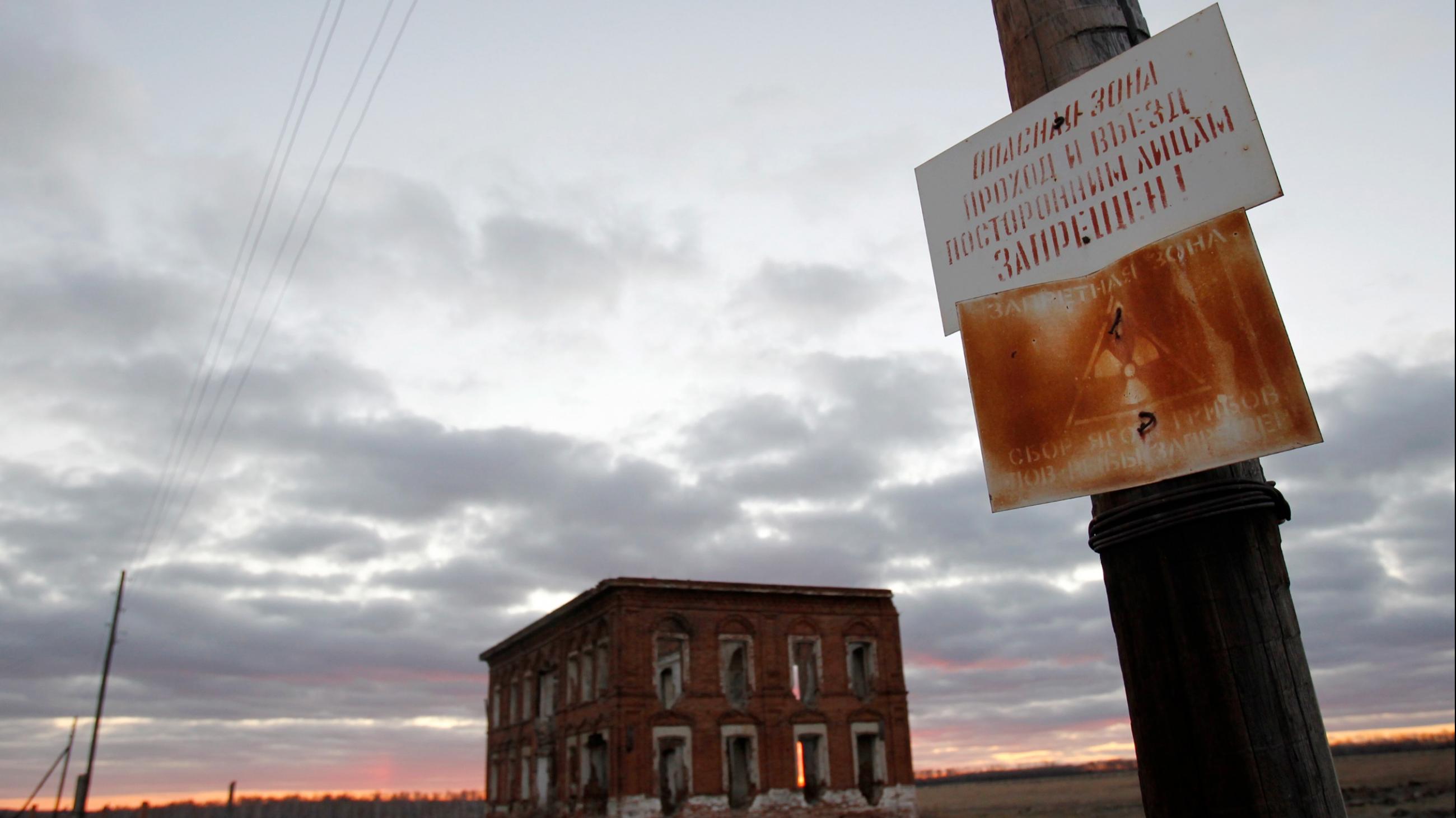 A rusted sign forbidding the gathering of mushrooms, picking berries and fishing is seen in front of an abandoned school which is silhouetted by a cloud-covered blue and orange sunset