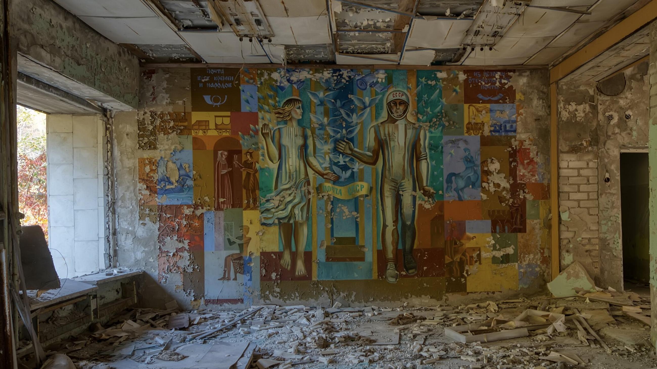 A chipping mural depicts a Soviet Kosmonaut and a woman of science on wall of a decaying building. The floor is littered with grey rubble and the ceiling tiles are cracked and falling. 