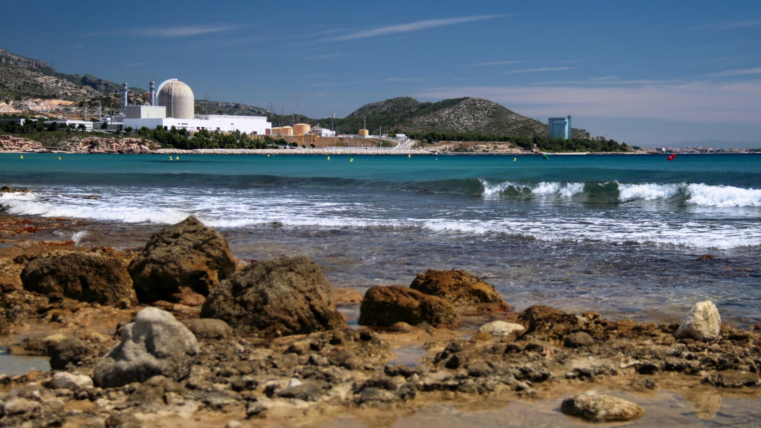 A view from a rocky beach on Spain's coastline. Across a crescent of turquoise ocean, waves cresting with white foam, can be seen the Vandellos II nuclear power plant sitting between green hills