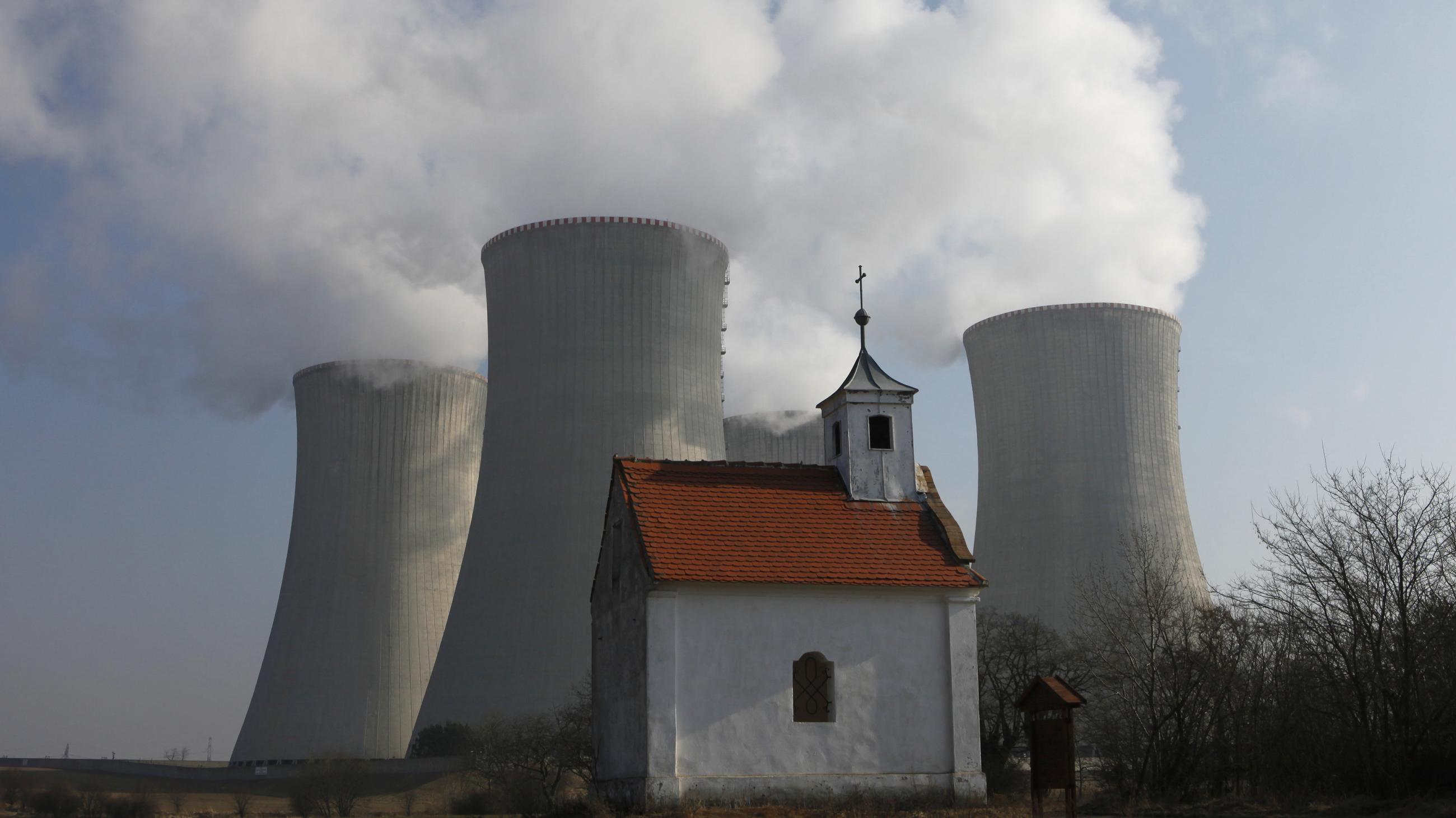 A small white chapel with a red roof stands in front of four concrete reactor towers, each billowing white smoke. 