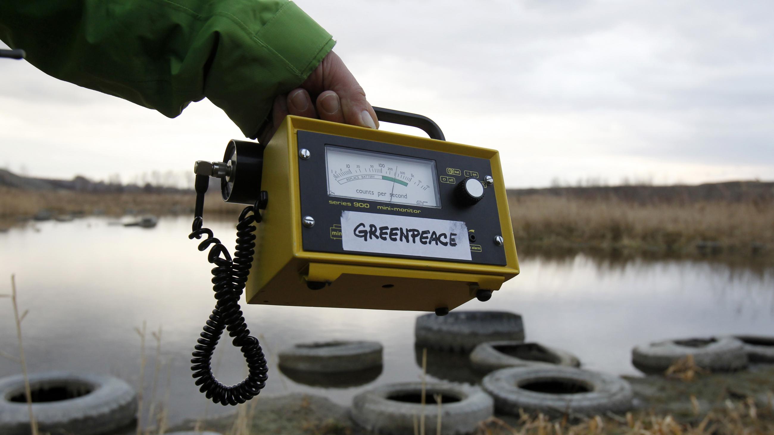 An extended arm in a green coat sleeve holds out a yellow Geiger counter, which shows a high level of radiation over the the banks of the Techa river, which is relatively still. Its banks are covered with old rubber tires and tall yellow grass