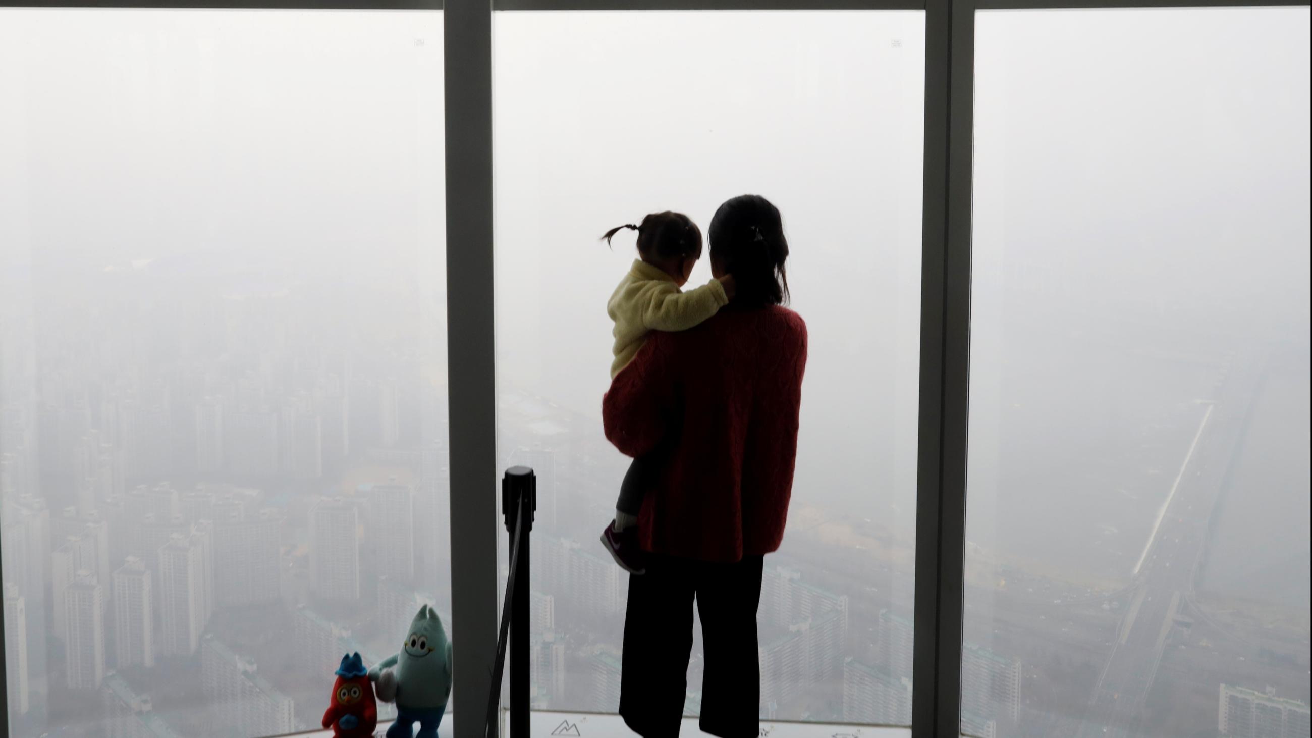 A woman holding her baby in her arms looks out a window at the city shrouded by a fine dust during a polluted day. 