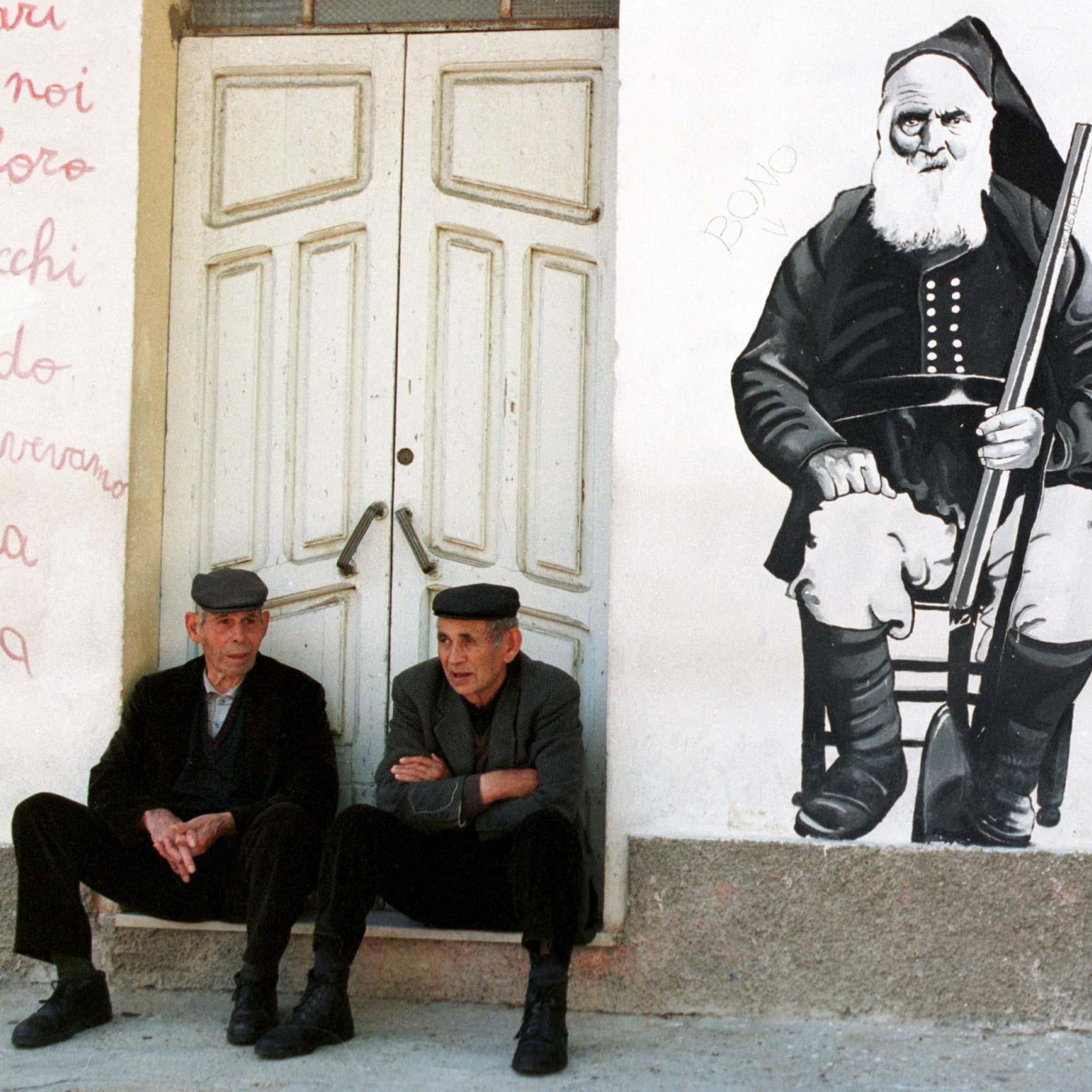 Two elderly men dressed in black suits with matching caps sit on a step in front of a black and white mural depicting an older man with a white beard wearing a military uniform