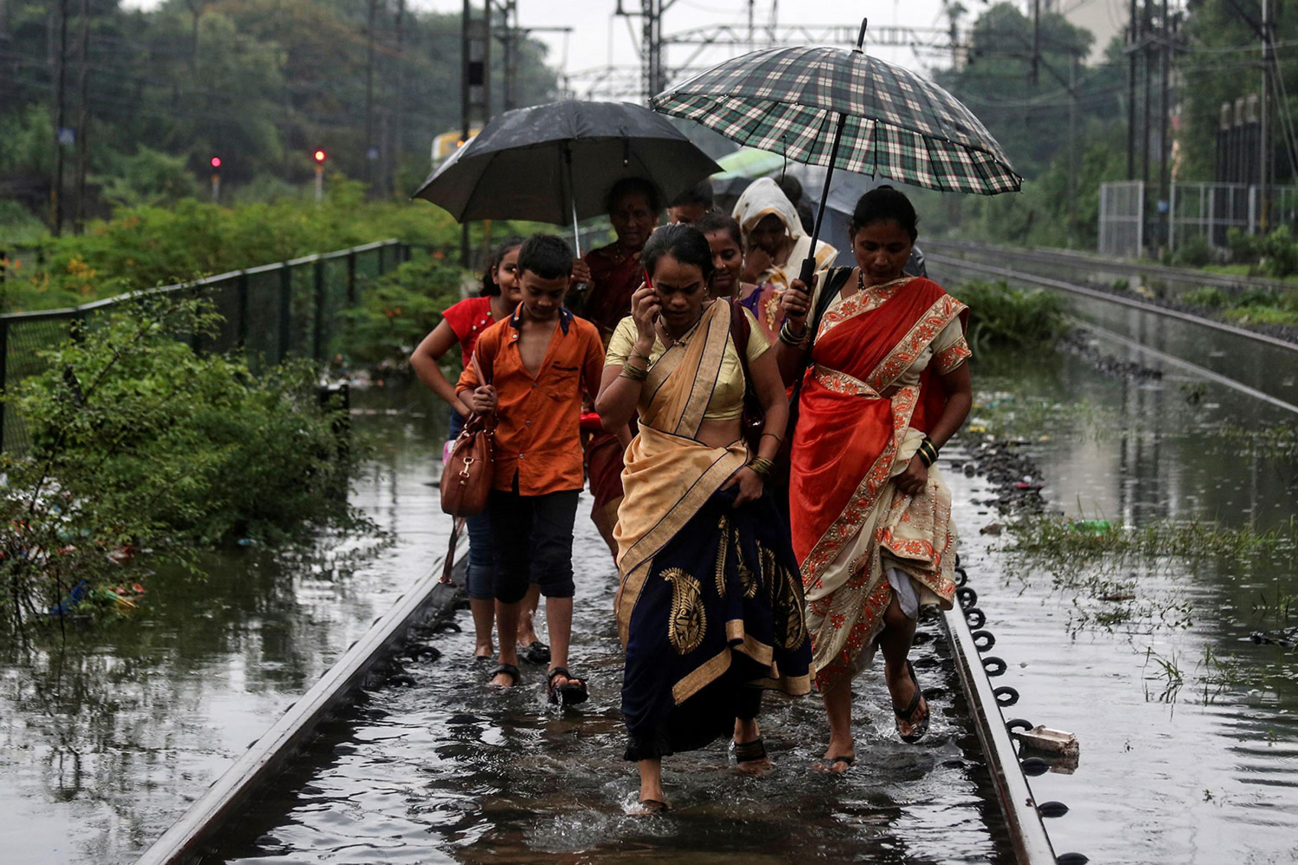 Commuters walk on waterlogged railway tracks after getting off a stalled train during heavy monsoon rains in Mumbai, India, on July 2, 2019. Several women and at least one boy are walking in a group. One of them is talking on a cell phone. Several are sharing umbrellas. The water, even on the train tracks, which are presumably slightly elevated, is up to their ankles. REUTERS/Francis Mascarenhas