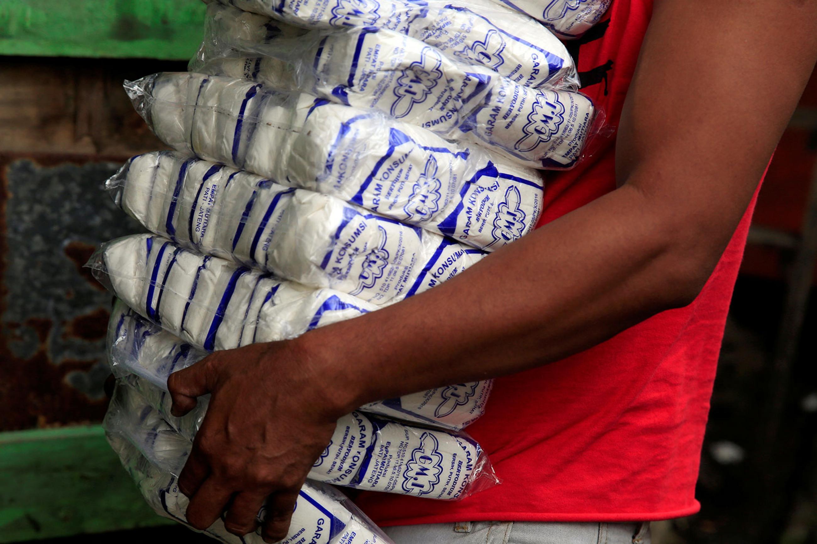 A worker carries packs of table salt at a warehouse in Jakarta, Indonesia, on March 14, 2018. Picture shows the torso of what appears to be a young man heavily burdened with more than a dozen large, one-pound bags of salt. He wears a red shirt and walks against a green backdrop and the bottom bag appears to be slipping out of his grasp. REUTERS/Beawiharta Beawiharta