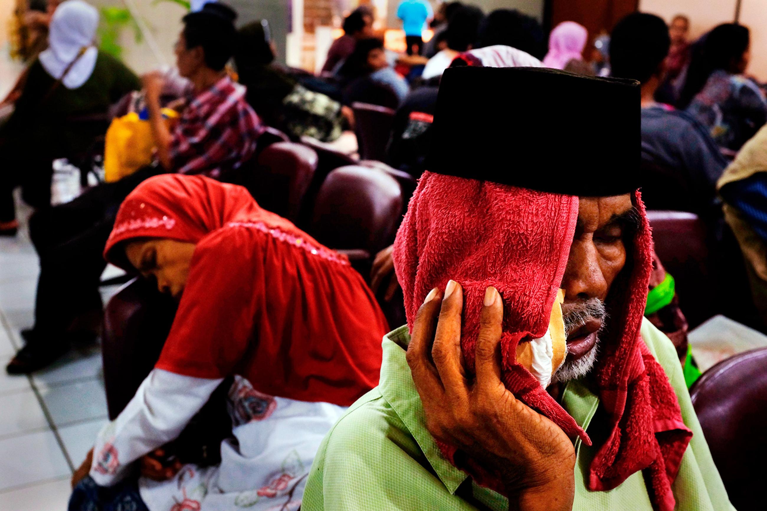 People waiting at Fatmawati hospital in Jakarta on May 17, 2013, after Indonesia announced plans to implement nationwide health care, with the aim to covering all Indonesians by the end of the decade. The picture shows a crowded waiting room with a man in the foreground with a red cloth draped over his head and holding what appears to be a bag of ice against his cheek. He looks like he is suffering. REUTERS/Beawiharta.