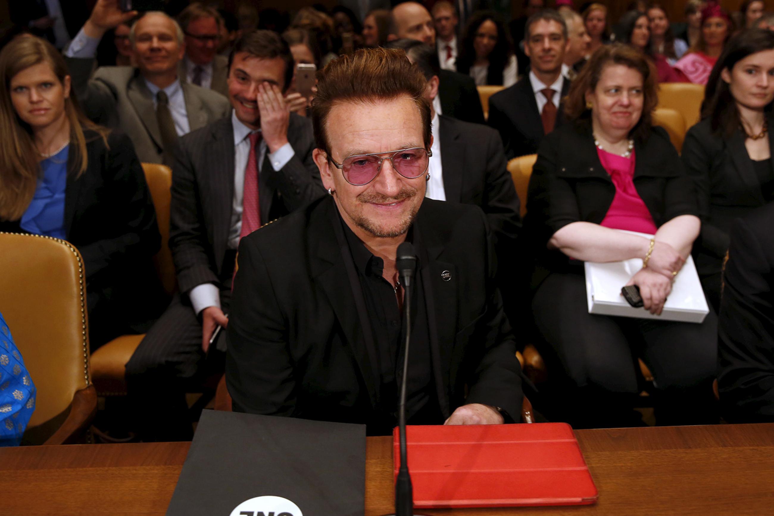 Famous singer and social activist Bono attends a Senate Appropriations State, Foreign Operations and Related Programs Subcommittee hearing on foreign assistance in Washington D.C. on April 12, 2016. The photo shows Bono at a long table with several rows of chairs behind him. The chairs are filled with people who appear to be laughing. The REUTERS/Yuri Gripas