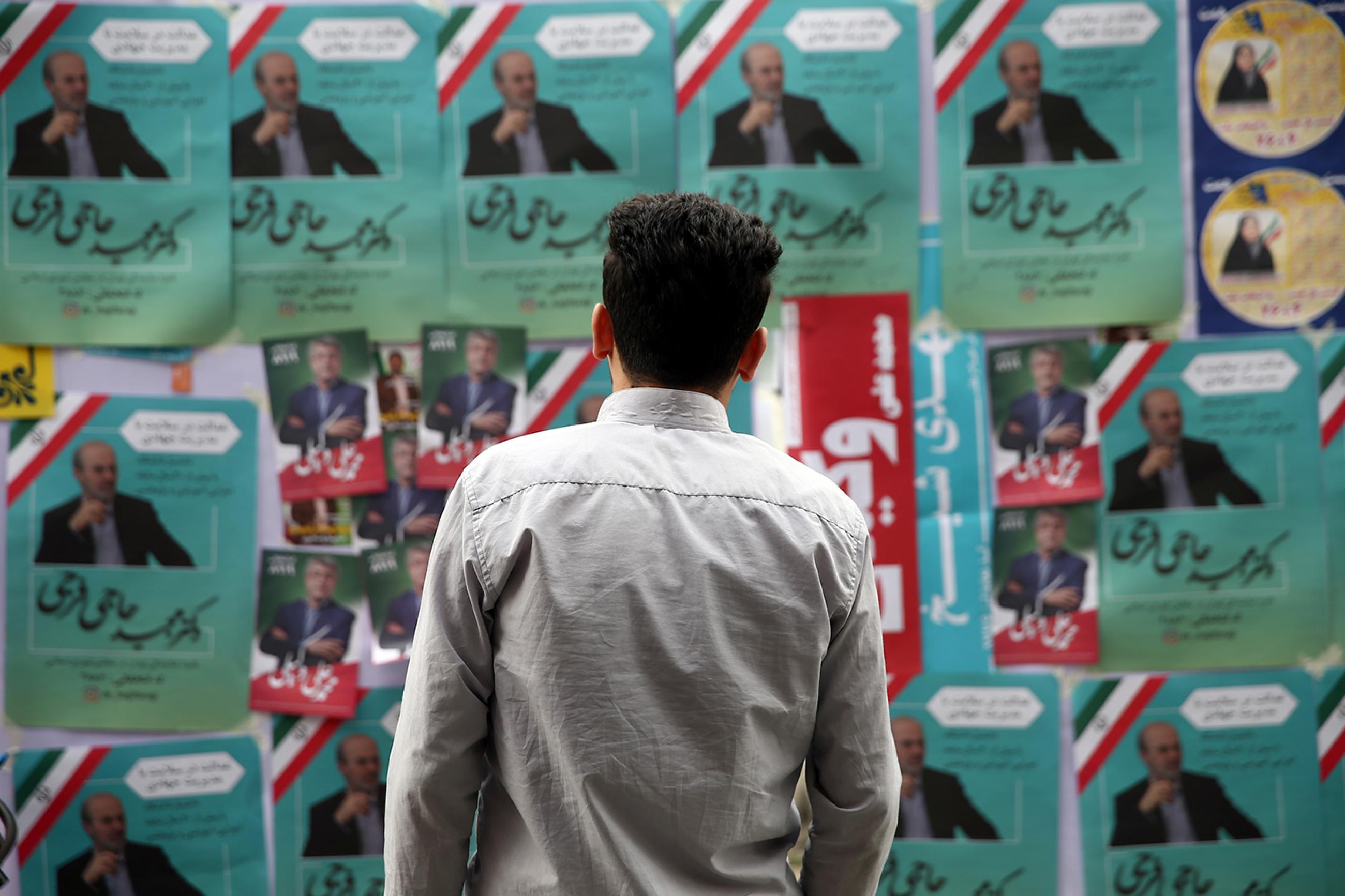 A man looks at parliamentary election campaign posters in Tehran, Iran on February 19, 2020. The photo shows the man standing in front of a board that is placarded with multiple political campaign posters, many of them identical and pasted over and over. The predominant colors are green, white, and red—like the Iranian flag. WANA/Nazanin Tabatabaee via REUTERS.
