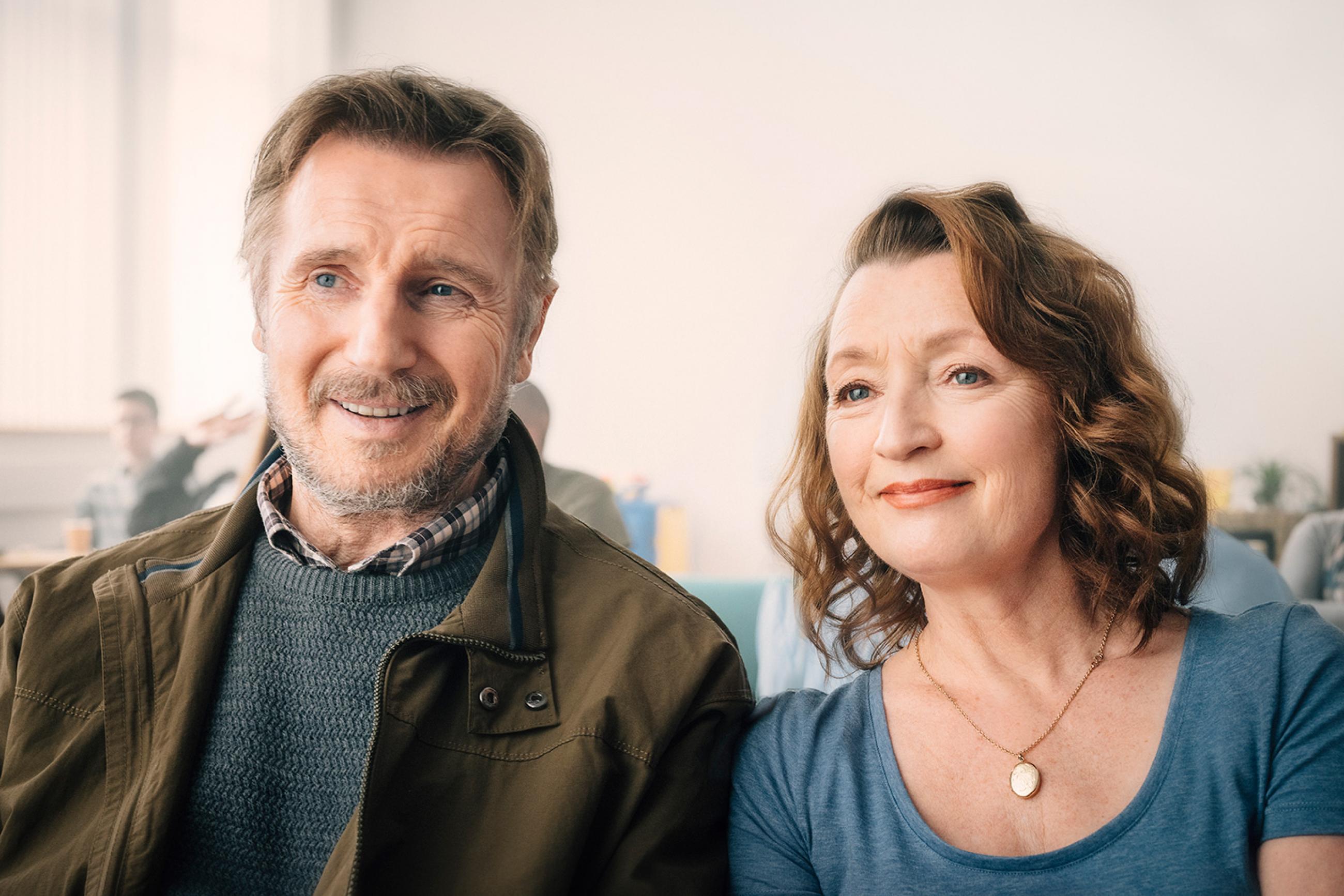 Liam Neeson and Lesley Manville give outstanding performances in the new film ORDINARY LOVE, from Bleecker Street, and they are helped by a powerful, semi-autobiographical script by Owen McCafferty. Image is of the two actors sitting together and smiling in a hospital waiting room. Credit: Aidan Monaghan/Bleecker Street