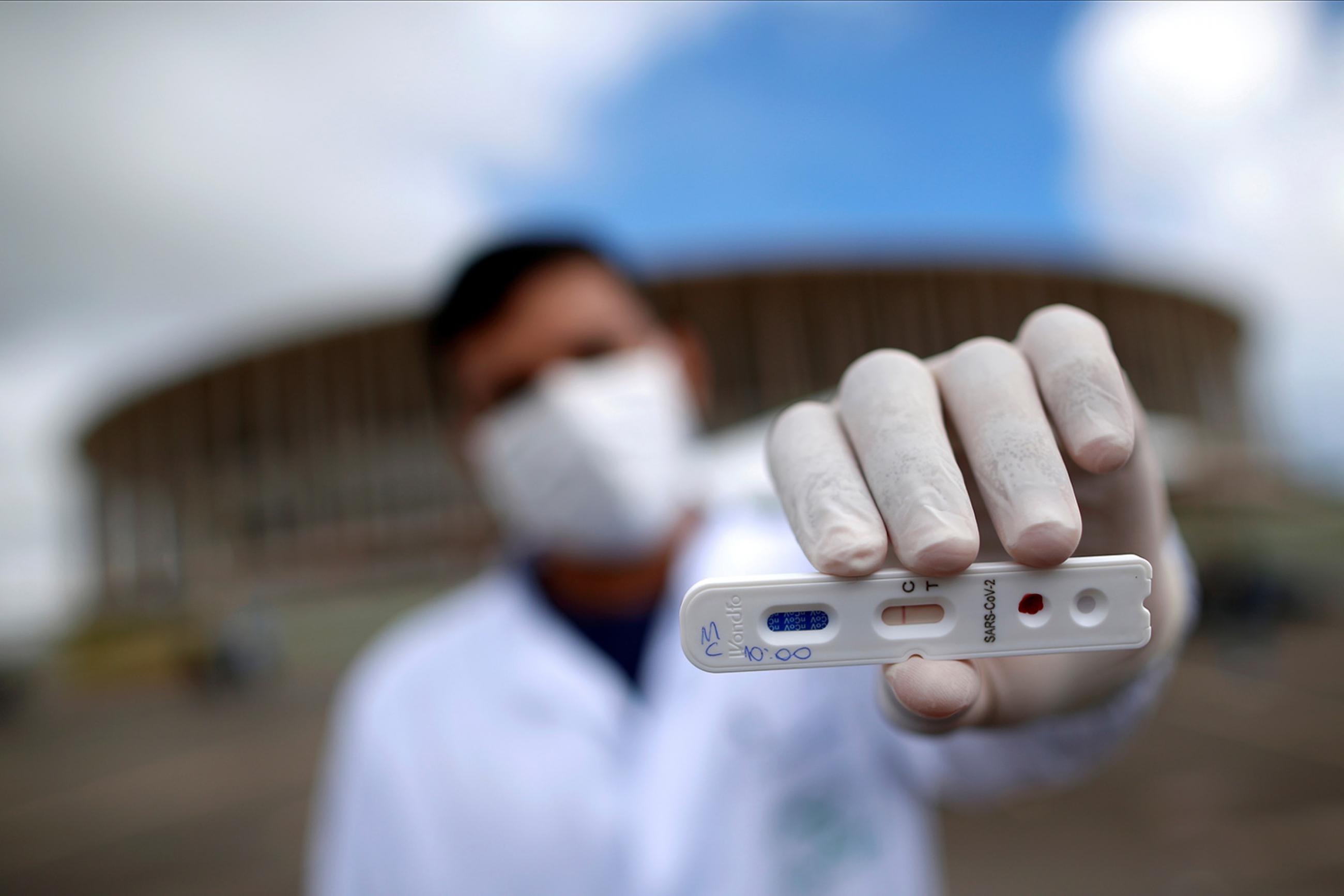 A medical professional shows a negative test for novel coronavirus in front of Mane Garrincha Stadium, amid the coronavirus disease (COVID-19) outbreak, in Brasilia, Brazil, on April 21, 2020. The photo shows a health worker holding a test at the camera with a stadium in the background. the photographer is using a telephoto/macro lens so that everything is blurred out in the shot, including the man, with the exception of the small plastic cassette-like test.  REUTERS/Ueslei Marcelino