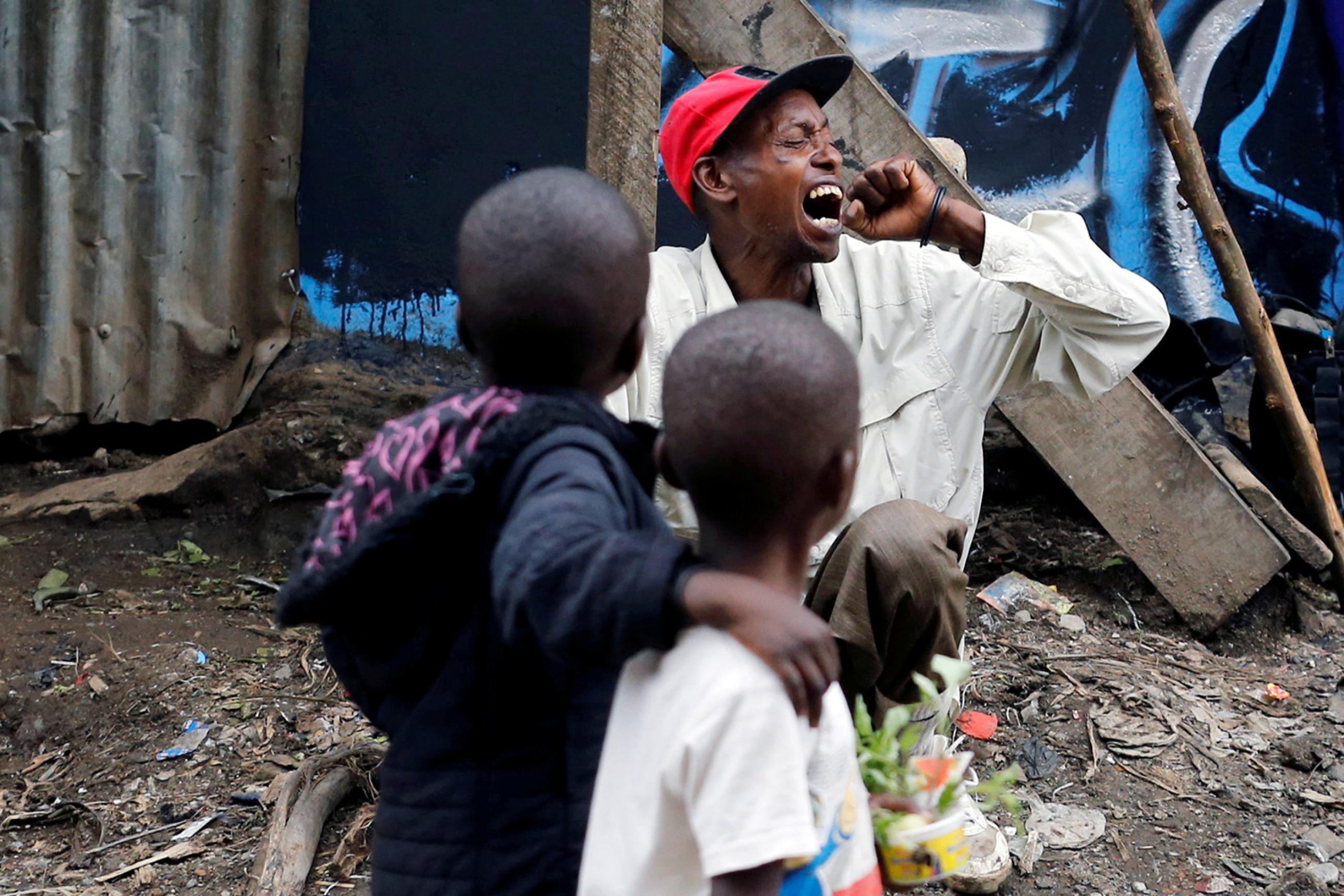 A man coughs as children walk past roadside, during the coronavirus pandemic in Nairobi, Kenya April 19, 2020. This is a powerful image of two children looking at a man who is coughing into his had with his head turned. REUTERS/Thomas Mukoya 