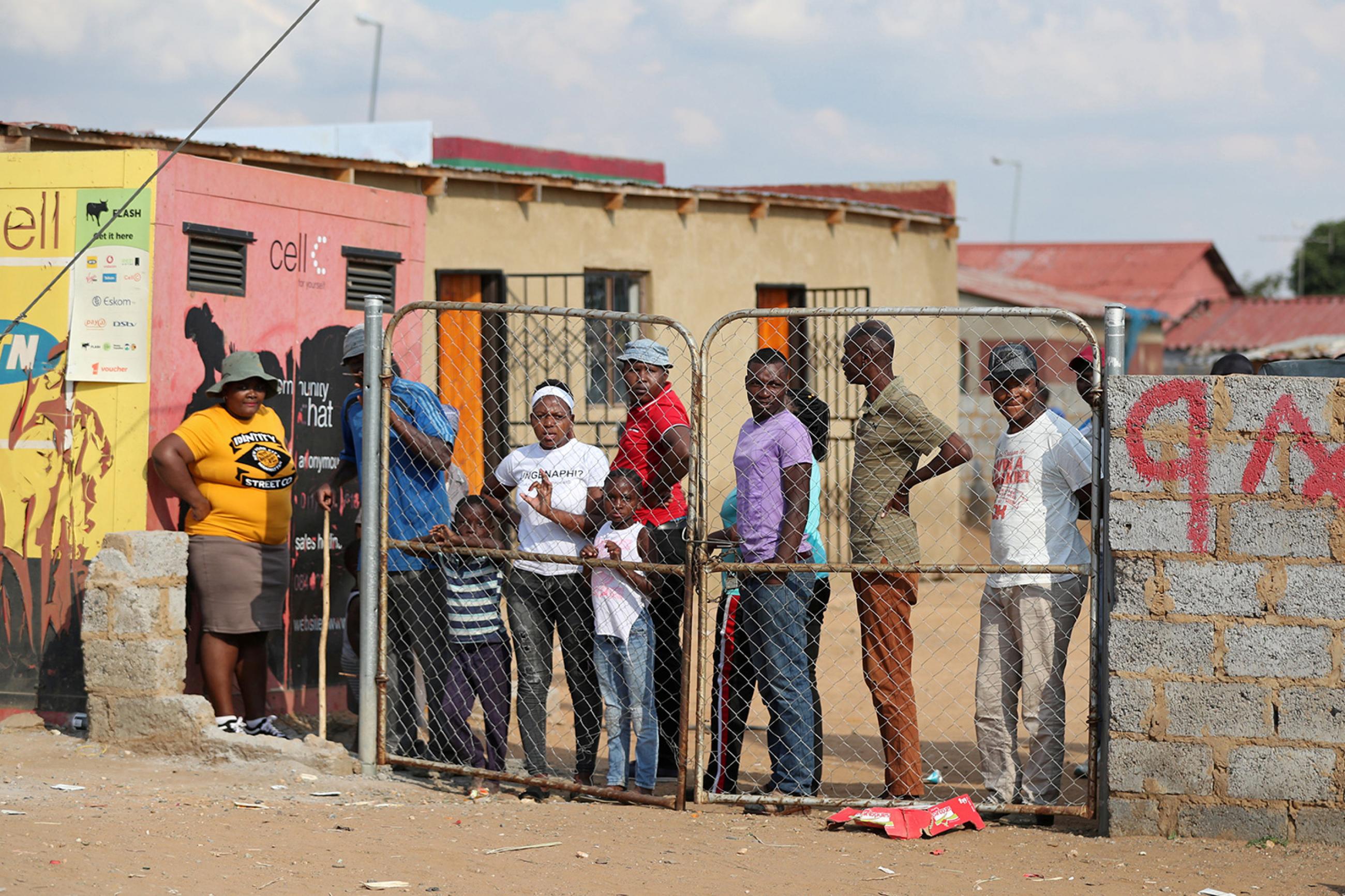 Residents look from behind a closed gate as members of the military patrol during a nationwide lockdown aimed at limiting the spread of novel coronavirus—in Soweto, South Africa on April 23, 2020. The photo shows several people congregated behind a closed fence though there is a very large gap between the fence and the adjacent wall. REUTERS/Siphiwe Sibeko 