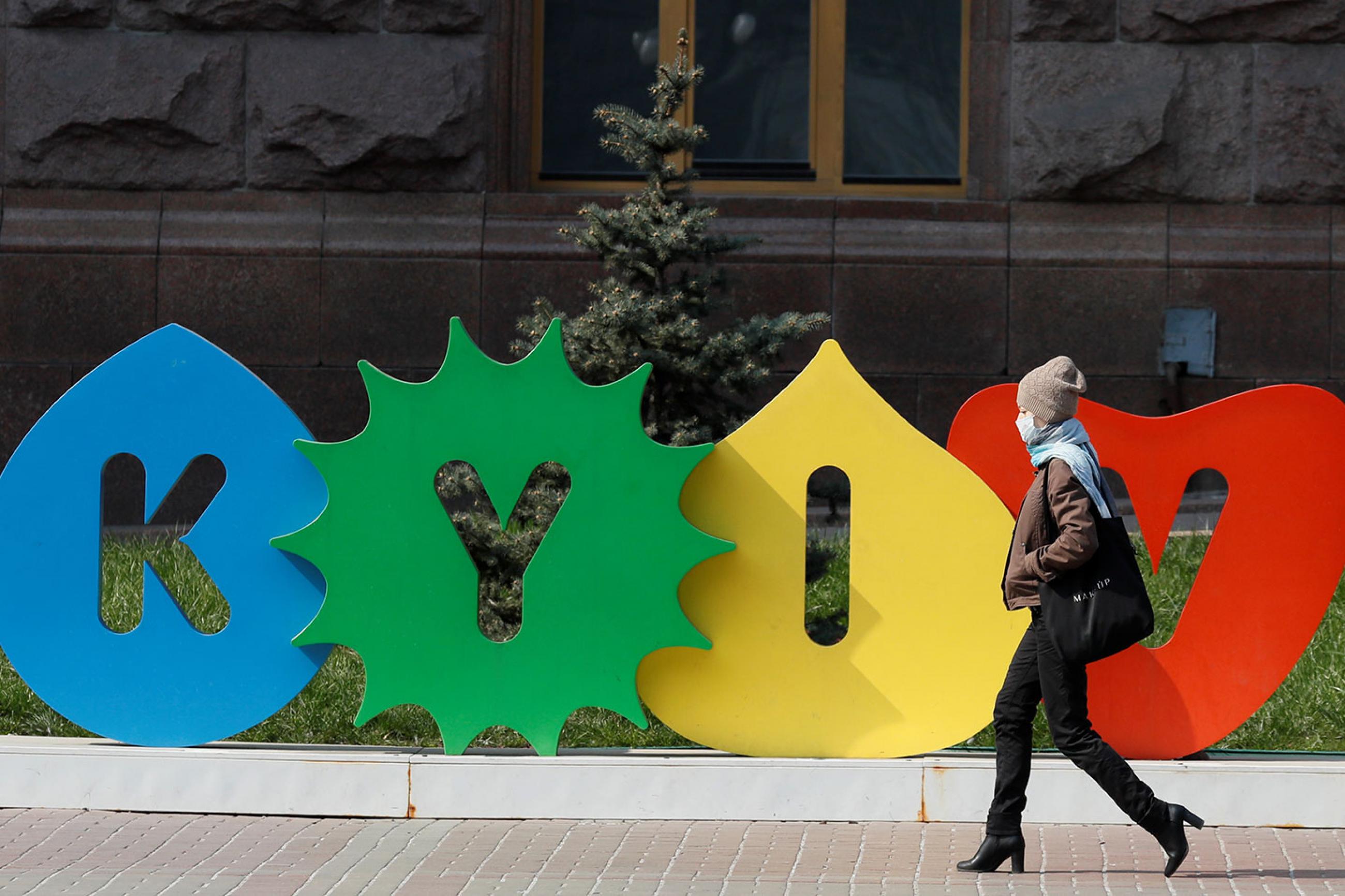 A woman wearing a mask during the coronavirus pandemic walks along the empty main street Khreshchatyk in central Kyiv, Ukraine on March 30, 2020—a colorful sign behind her bears the name of the city. This is a striking photo with a stylish woman walking in front of a sign with the city's name. REUTERS/Gleb Garanich