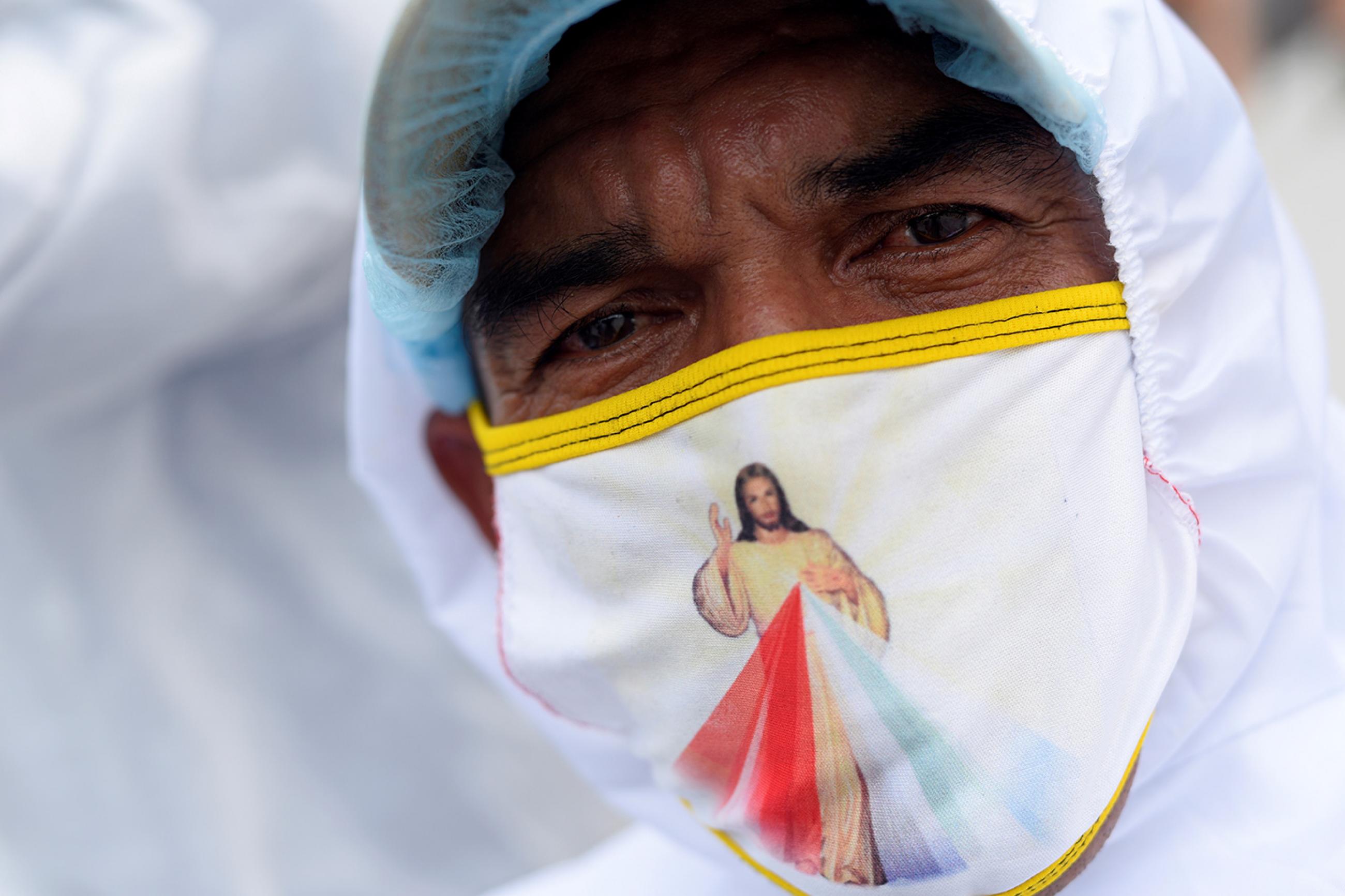 A funeral worker wears a face mask with an illustration of Jesus, outside Los Ceibos hospital amid the outbreak of the coronavirus disease (COVID-19), in Guayaquil, Ecuador, on April 15, 2020. The photo shows the worker in protective head covering and mask. on his mask is an image of Jesus with a rainbow colors on his robe. REUTERS/Santiago Arcos