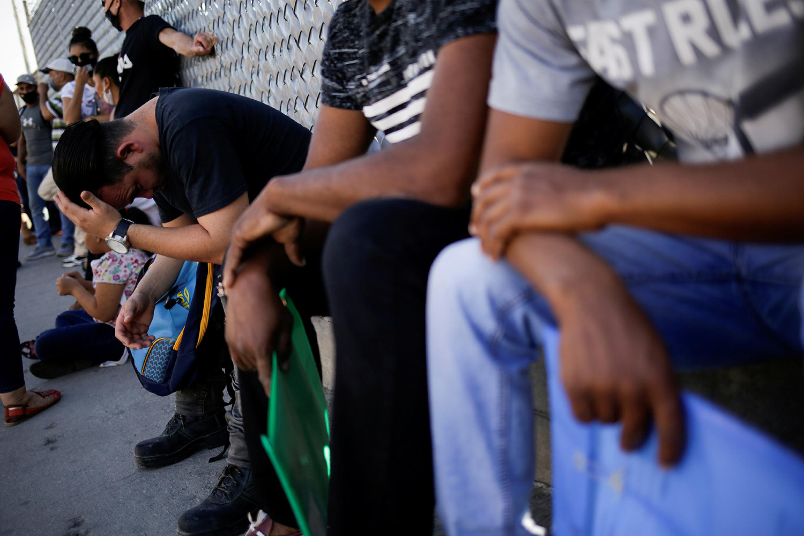 Migrants in the "Remain in Mexico" program queue in Juarez, Mexico, on July 8, 2020 to renew their permission to stay legally in Mexico to wait for their immigration hearing in the United States. This is a powerful image showing people lined up seated with their backs to a chain-link fence covered in barbed wire. Some of them hold packets of papers. One of them has his head resting in his hands.  REUTERS/Jose Luis Gonzalez