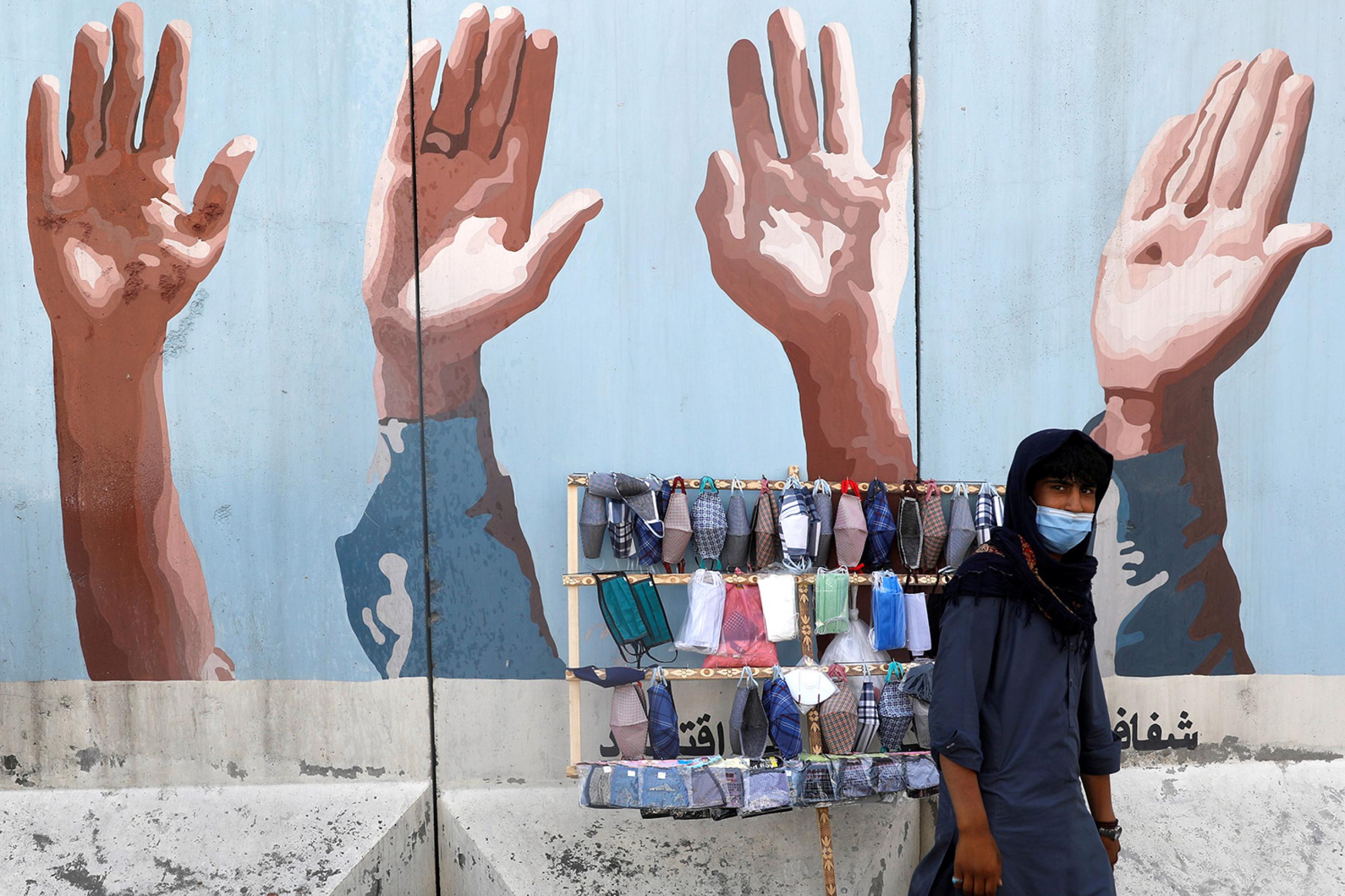 An Afghan man sells protective face masks during the coronavirus disease (COVID-19) outbreak in Kabul, Afghanistan June 18, 2020. The photo shows a man in front of a small cart with masks on display. in the background is a mural of hands raised. REUTERS/Mohammad Ismail