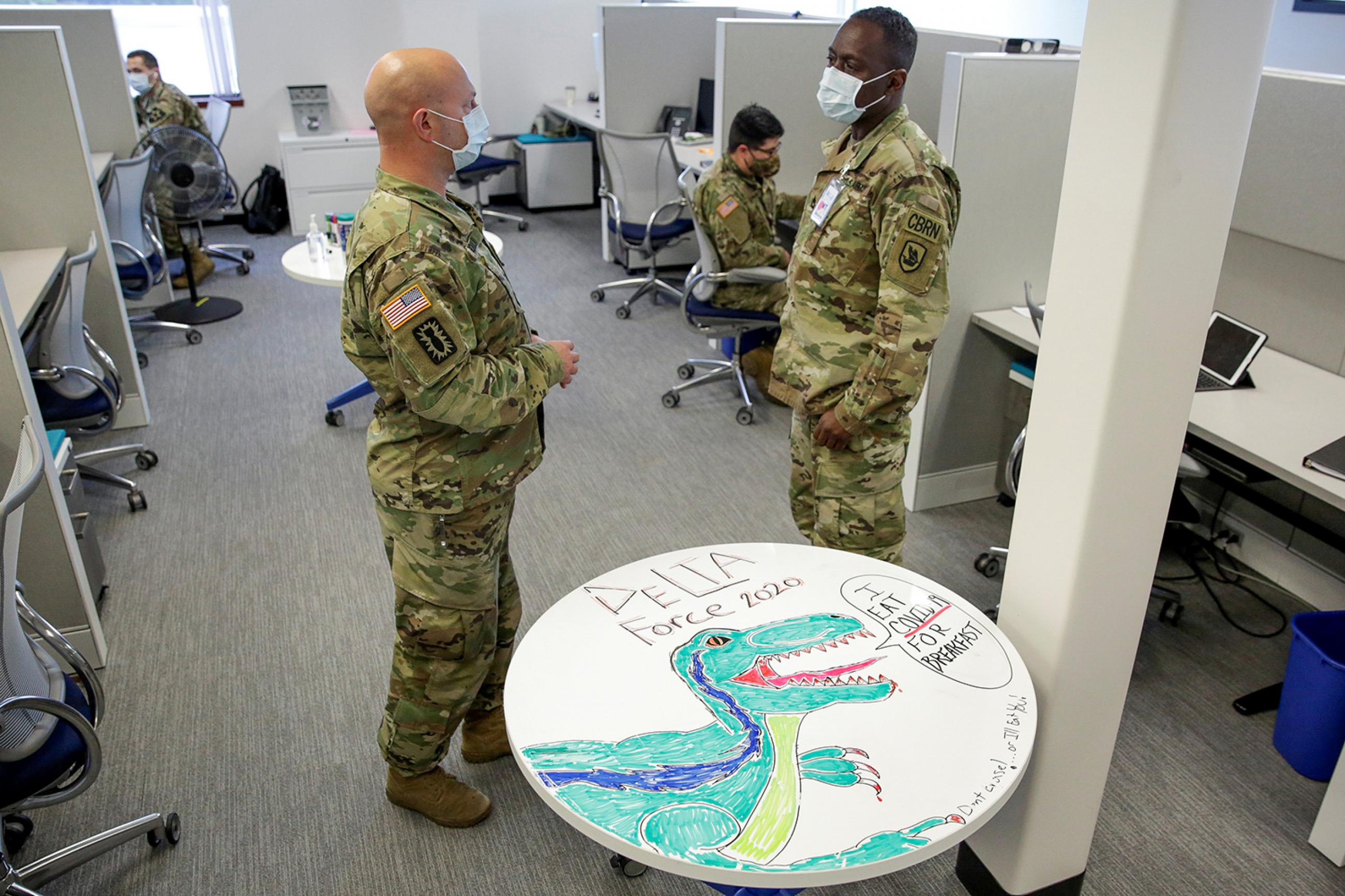 A militarized approach to contact tracing is shown here as National Guard members at the Washington State Department of Health support contact tracing efforts in Tumwater, Washington—on May 20, 2020. The photo shows the guard members standing around a cubicle space talking. A table in the foreground is decorated with a drawing of a dinosaur and emblazoned with the words "I eat COVID-19 for breakfast. REUTERS/Jason Redmond