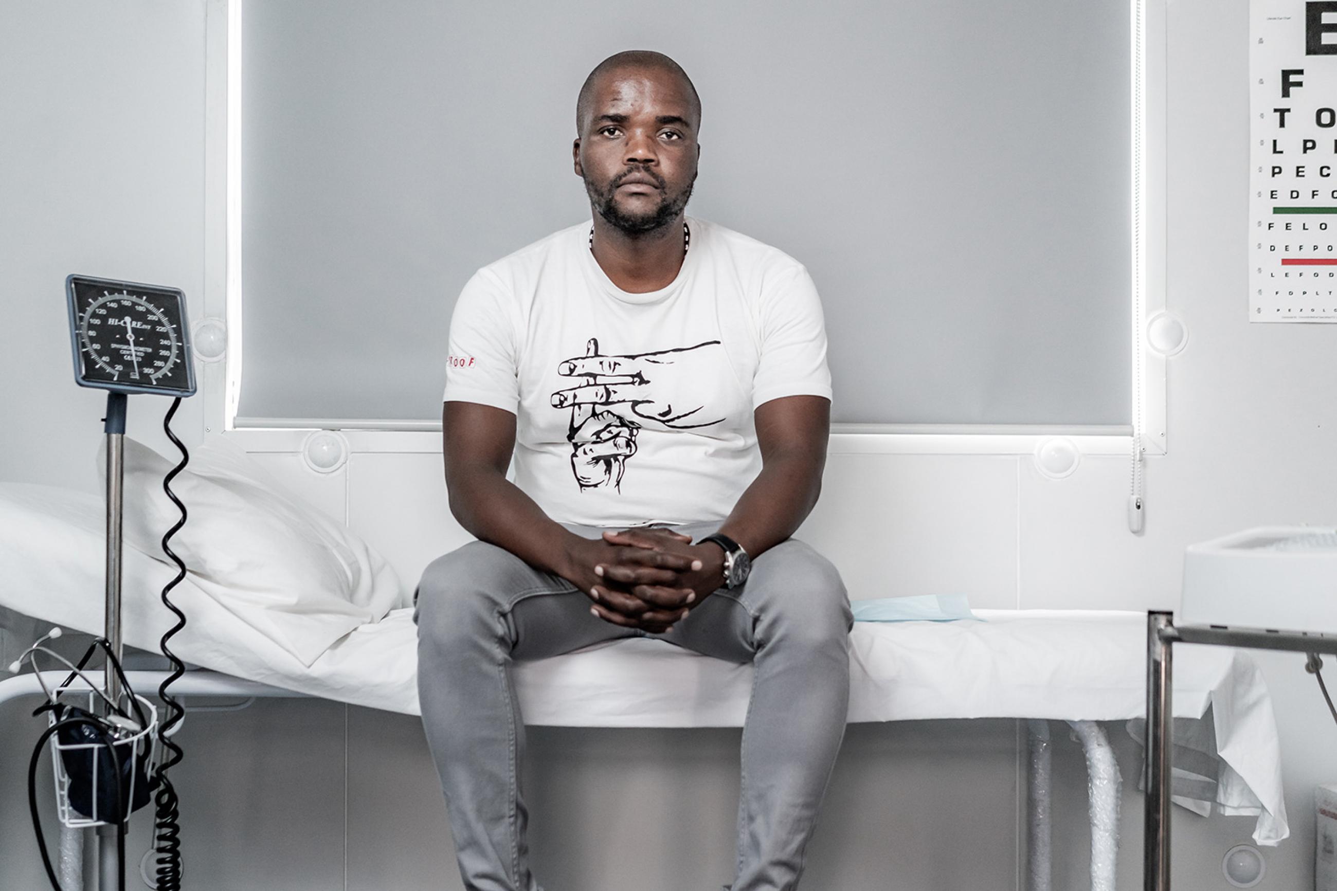Goodman Makanda, who's involved in efforts to make tuberculosis and COVID-19 testing more acceptable and accessible for South African men, in Khayelitsha, Cape Town, South Africa, on March 24th 2019. The photo shows a man sitting in a clinic with various monitors and equipment. An eye exam is visible on the wall. MSF/Jelle Krings