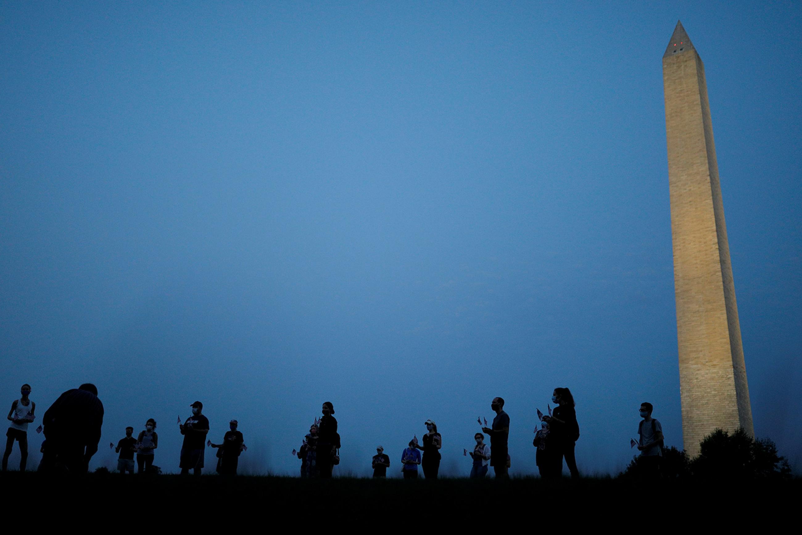 They are informally honored—but are they officially counted? Activists stand at sunrise to memorialize victims of the coronavirus, near the Washington Monument in Washington, DC on August 27, 2020. The photo shows the monument in the thin morning lite with a silhouette line of people standing in the foreground. REUTERS/Tom Brenner