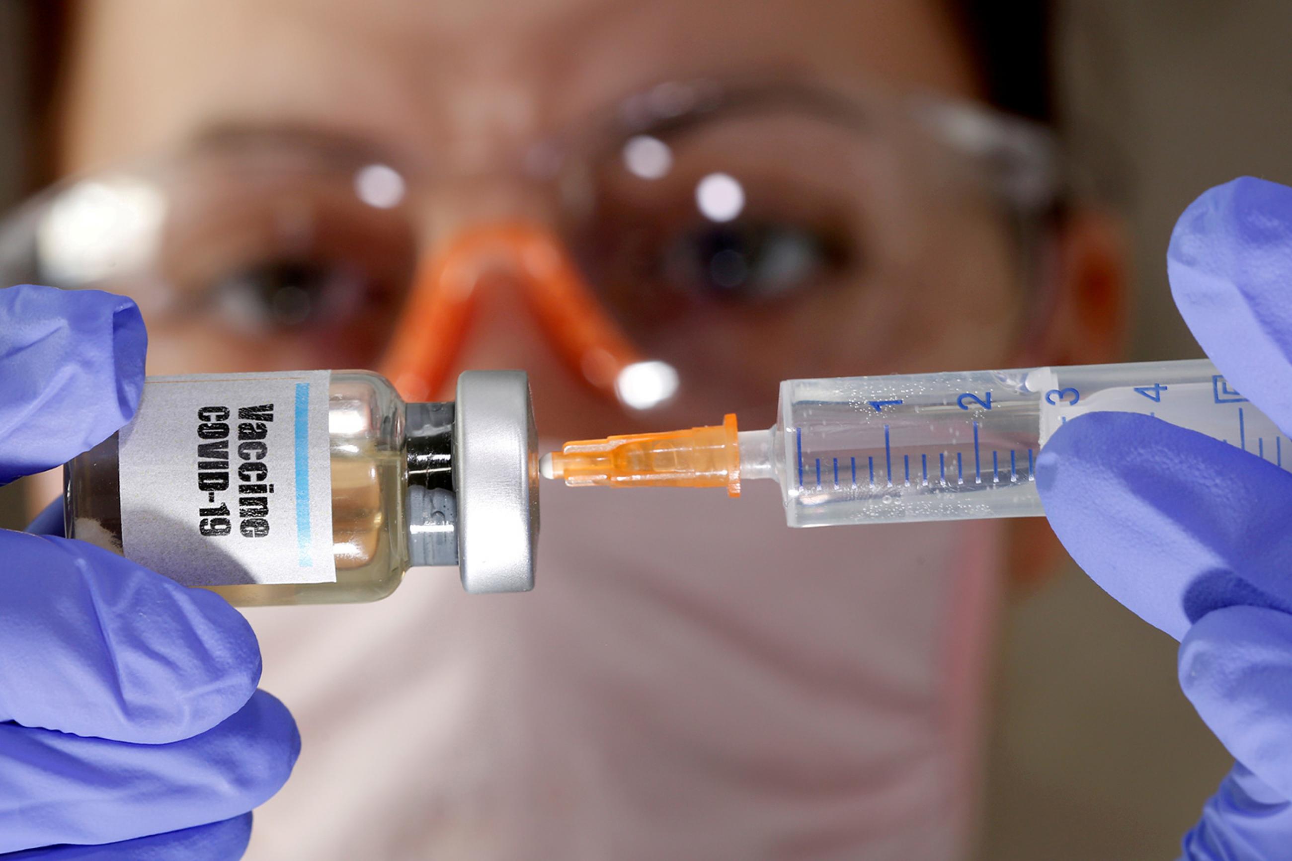 A woman holds a small bottle labeled with a "Vaccine COVID-19" sticker and a medical syringe in this illustration taken on April 10, 2020, in Bosnia. The photo shows a woman holding a small vial to her face. She is wearing a mask and gloves and the vial reads, COVID-19. REUTERS/Dado Ruvic
