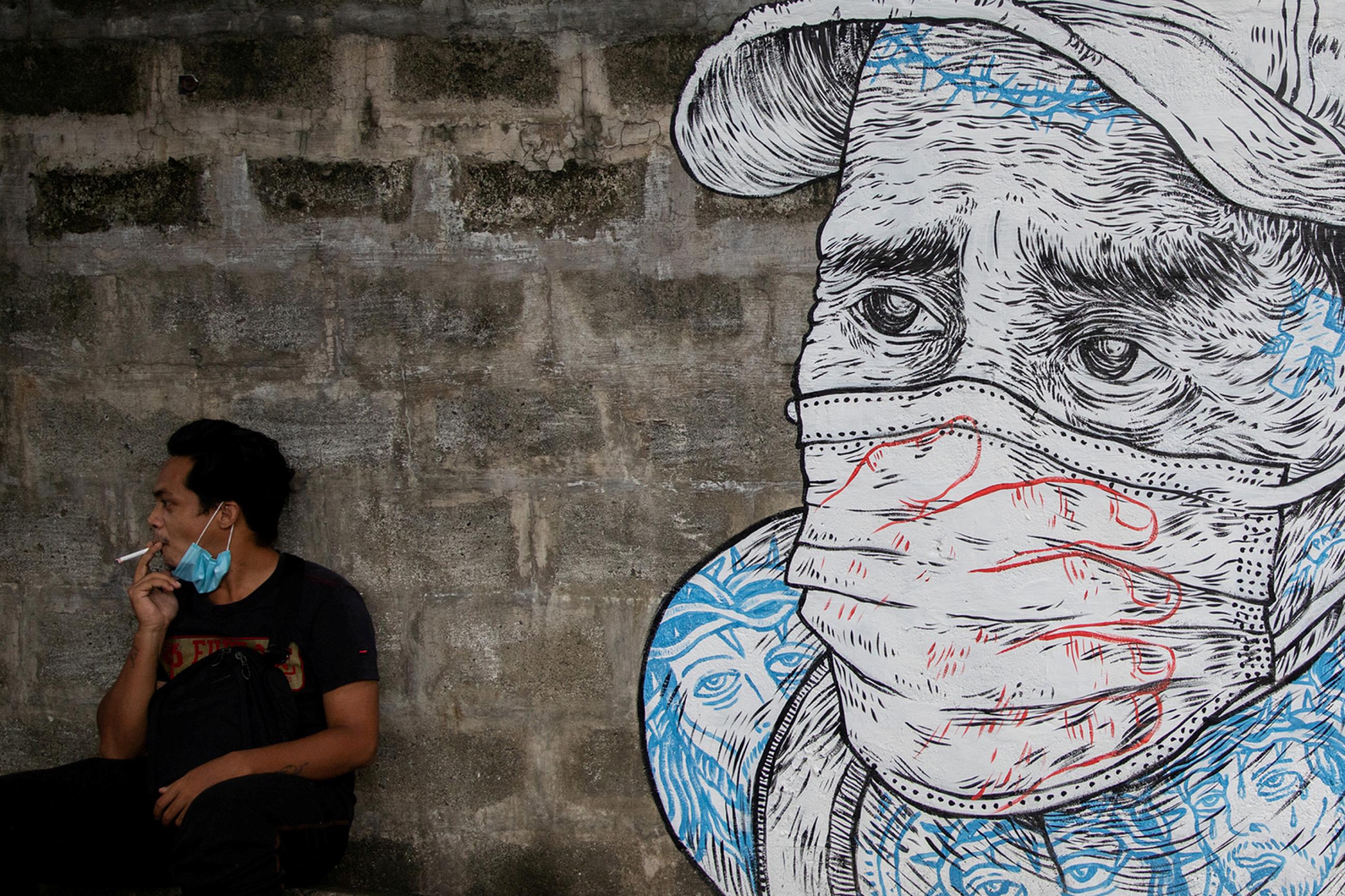A man smokes a cigarette next to a mural of a man wearing a protective mask amid the coronavirus disease (COVID-19) outbreak in Quezon City, Metro Manila, Philippines, July 30, 2020. The photo shows a man wearing a mask pulled down around his chin smoking. Next to him is a mural of a worried-looking man in a mask. REUTERS/Eloisa Lopez